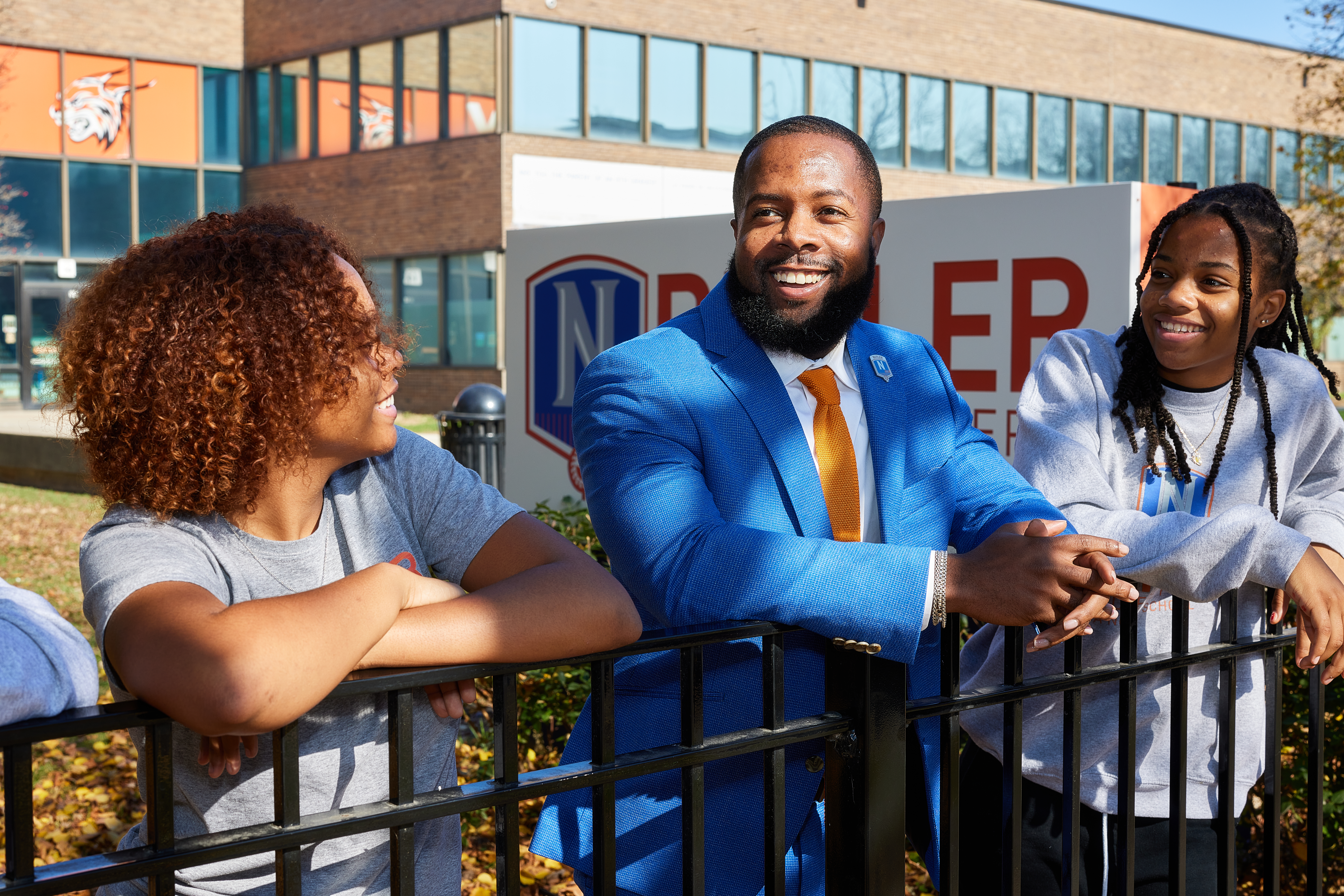 Photo shows Principal Brian Riddick talking with two Butler College Prep students outside the school building. They are all smiling and looking at him, like they are having an engaging conversation.