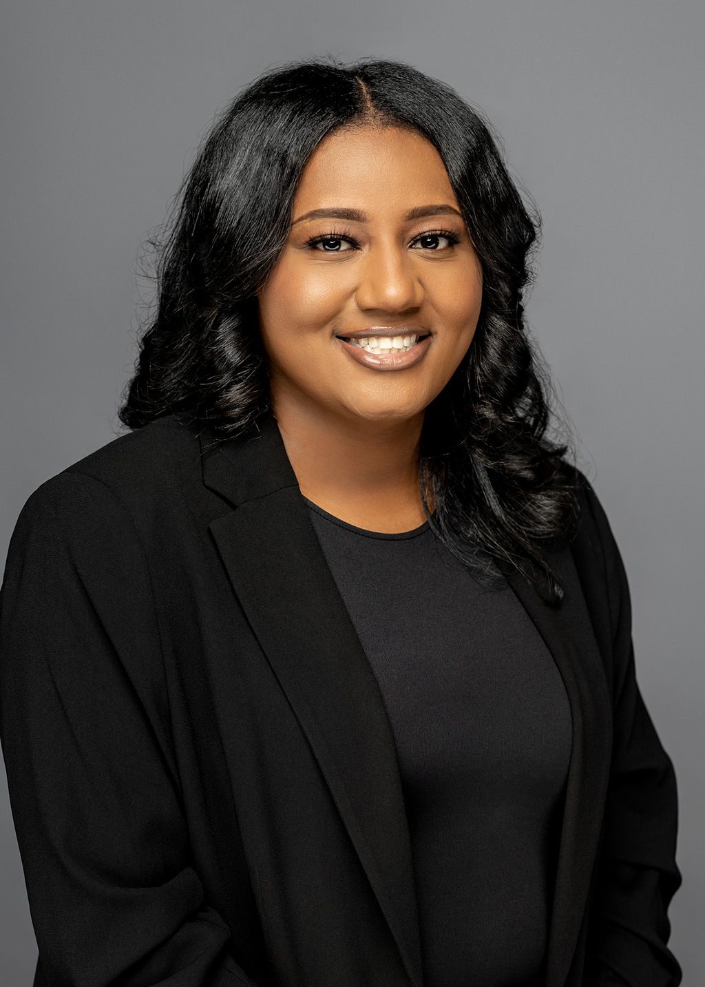 This photo is a professional headshot of Cierra Howard. She is a Black woman with medium-length straight black hair in loose curls. She is wearing a black blouse with a black blazer on top of it. She is smiling and her arms are relaxed to her sides with her hands slightly crossed in front.