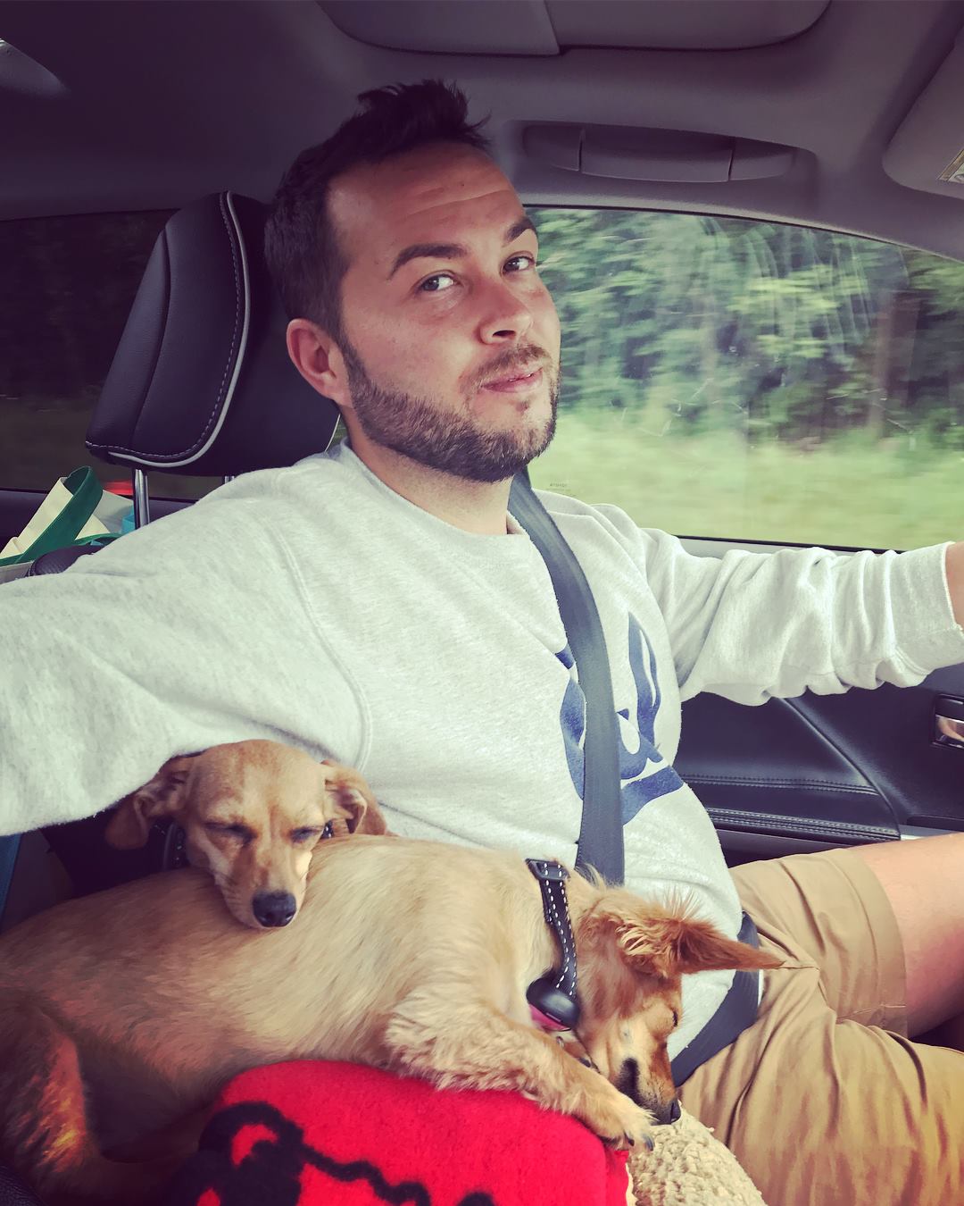 Photo shows Chase Johnson-Espinoza, principal of Muchin College Prep, driving in a car and holding two small dogs who are asleep on top of each other.