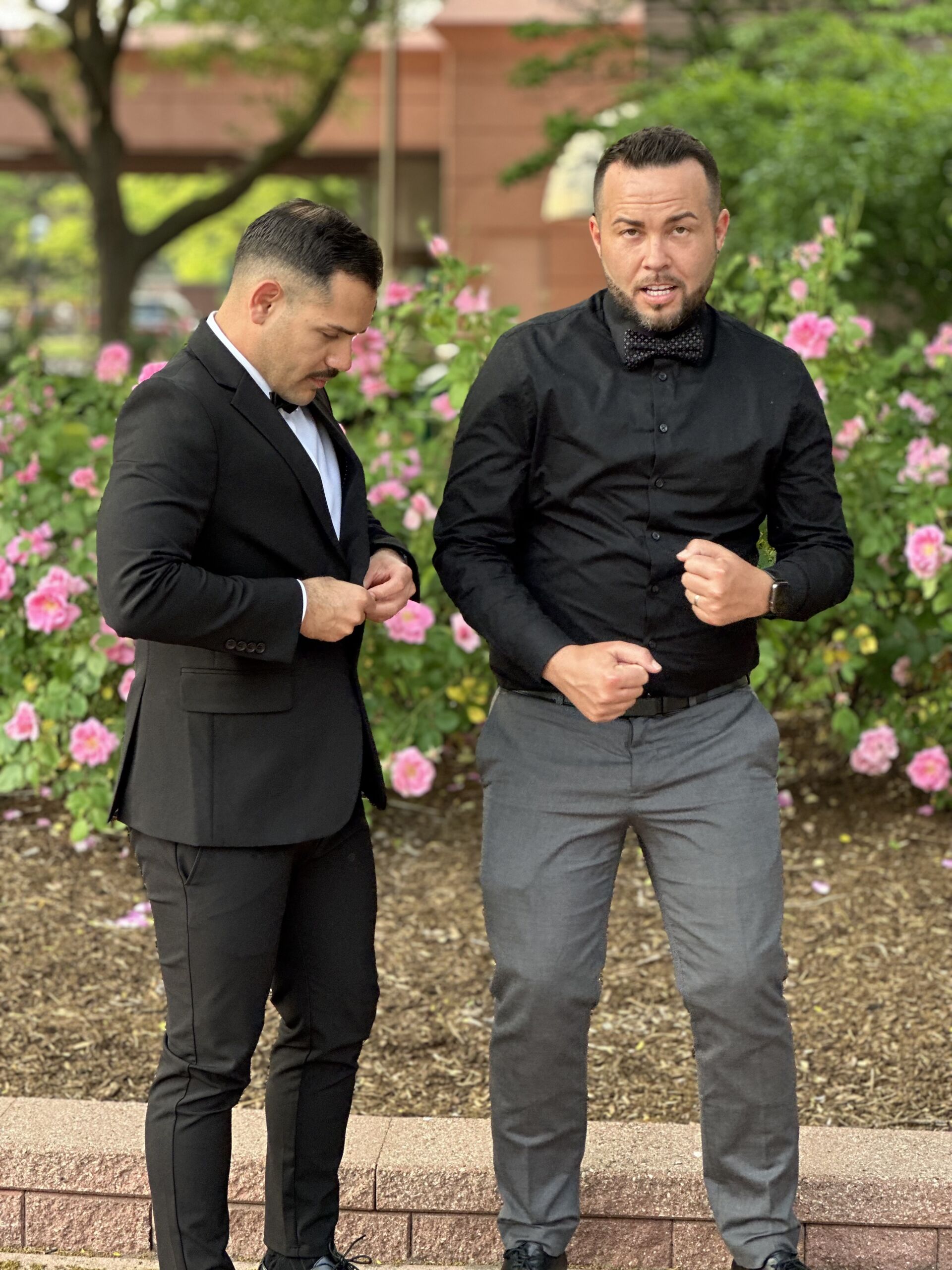 Photo shows a picture of Chase Johnson-Espinoza at his wedding. He is standing, looking like he's ready to dance, in front of a patch of flowers outside, alongside another person in formal wear. He is wearing a black long-sleeve button up with gray slacks and a black bowtie with white dots.
