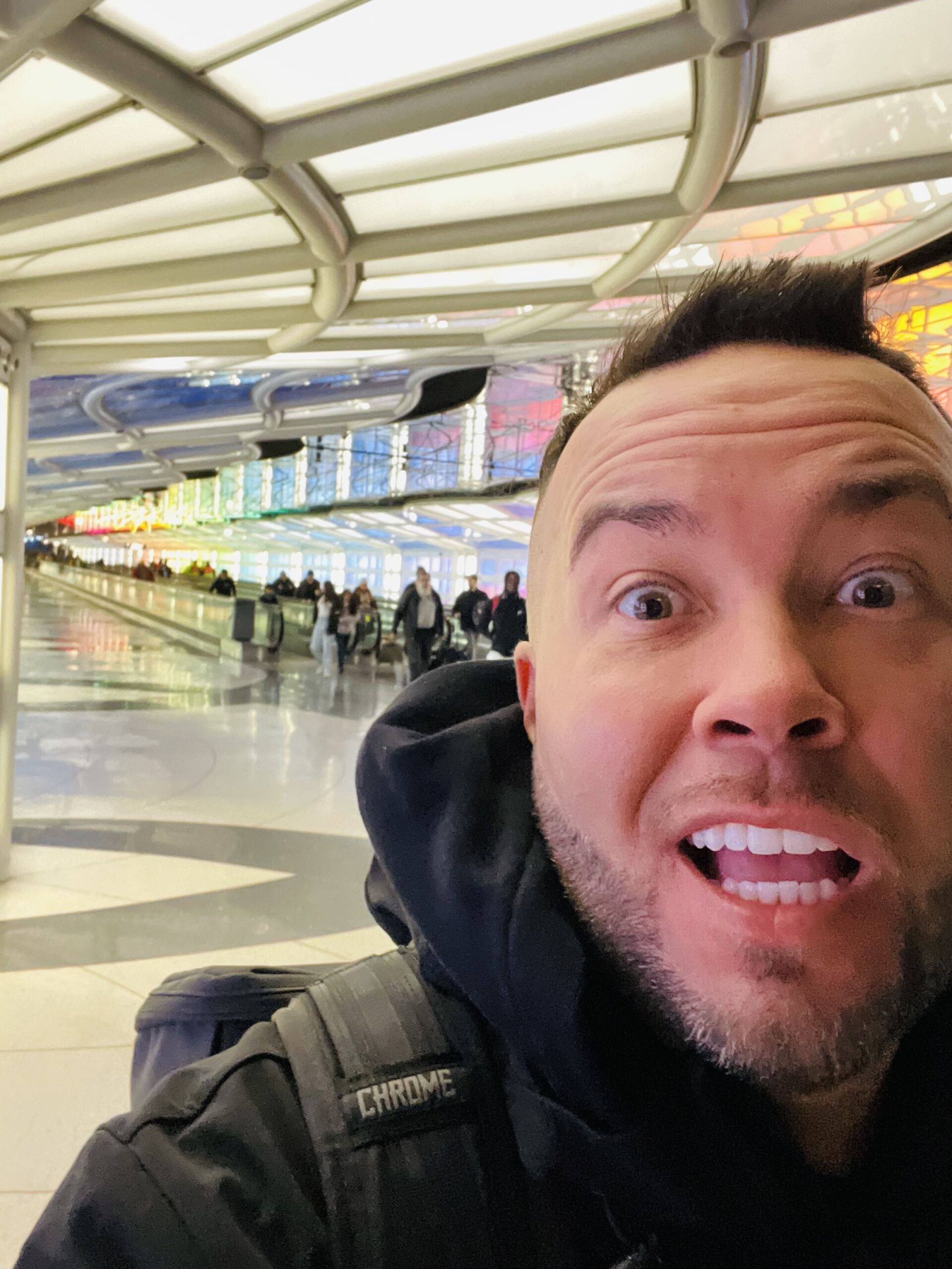 Photo is a humorous selfie of Chase Johnson-Espinoza in an airport. He has an excited and funny facial expression on, with his eyebrows raised high and his mouth and eyes wide open.