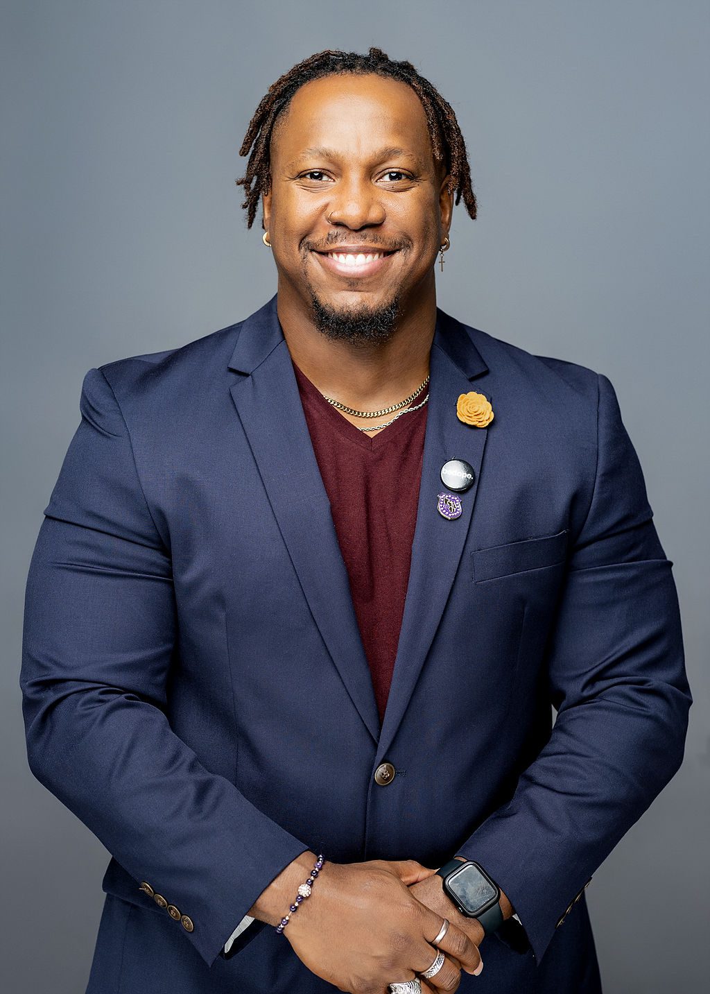 This photo is a professional headshot of Dyryl Burnett. He is a Black man with short dark brown locs. He is wearing a maroon t-shirt with a dark blue blazer closed over the top of it. On the left lapel of his blazer, there are three pins: a yellow flower, a black-and-white pin that says "be dope", and a Omega Psi Phi Fraternity crest. He is wearing two simple chains around his neck, one silver and one gold, and has two gold earrings on -- one of the earrings is a cross. Lastly, on his wrists, he is wearing a black smartwatch and a simple beaded bracelet. He is smiling and his arms are relaxed to his side with his hands clasped in front of his waist.