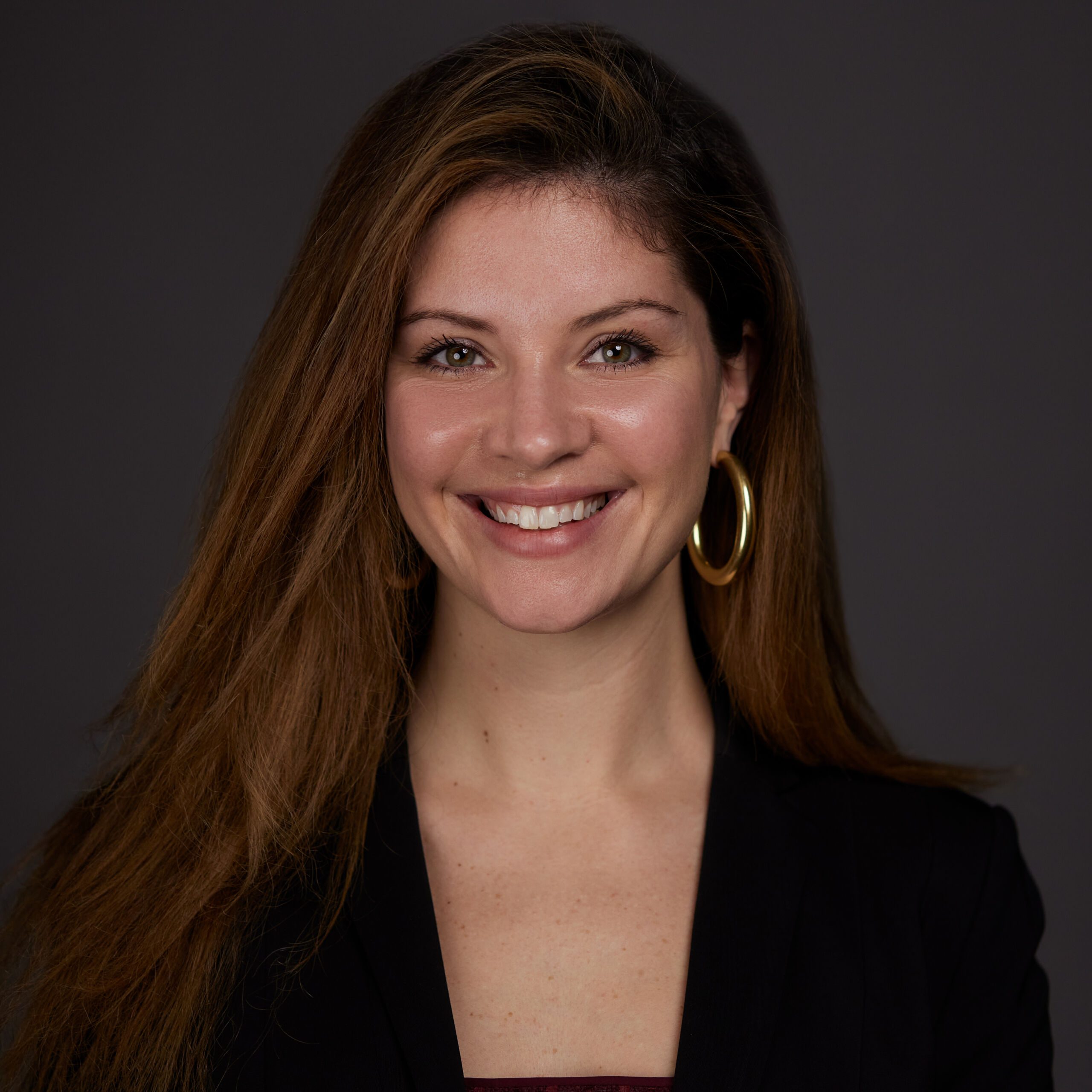 Photo shows a professional headshot of Jessie Weingartner, principal of Golder College Prep. She is a white woman with long straight brown hair. She is smiling and wearing a black long sleeve blouse and gold hoop earrings. The background is a solid gray color.