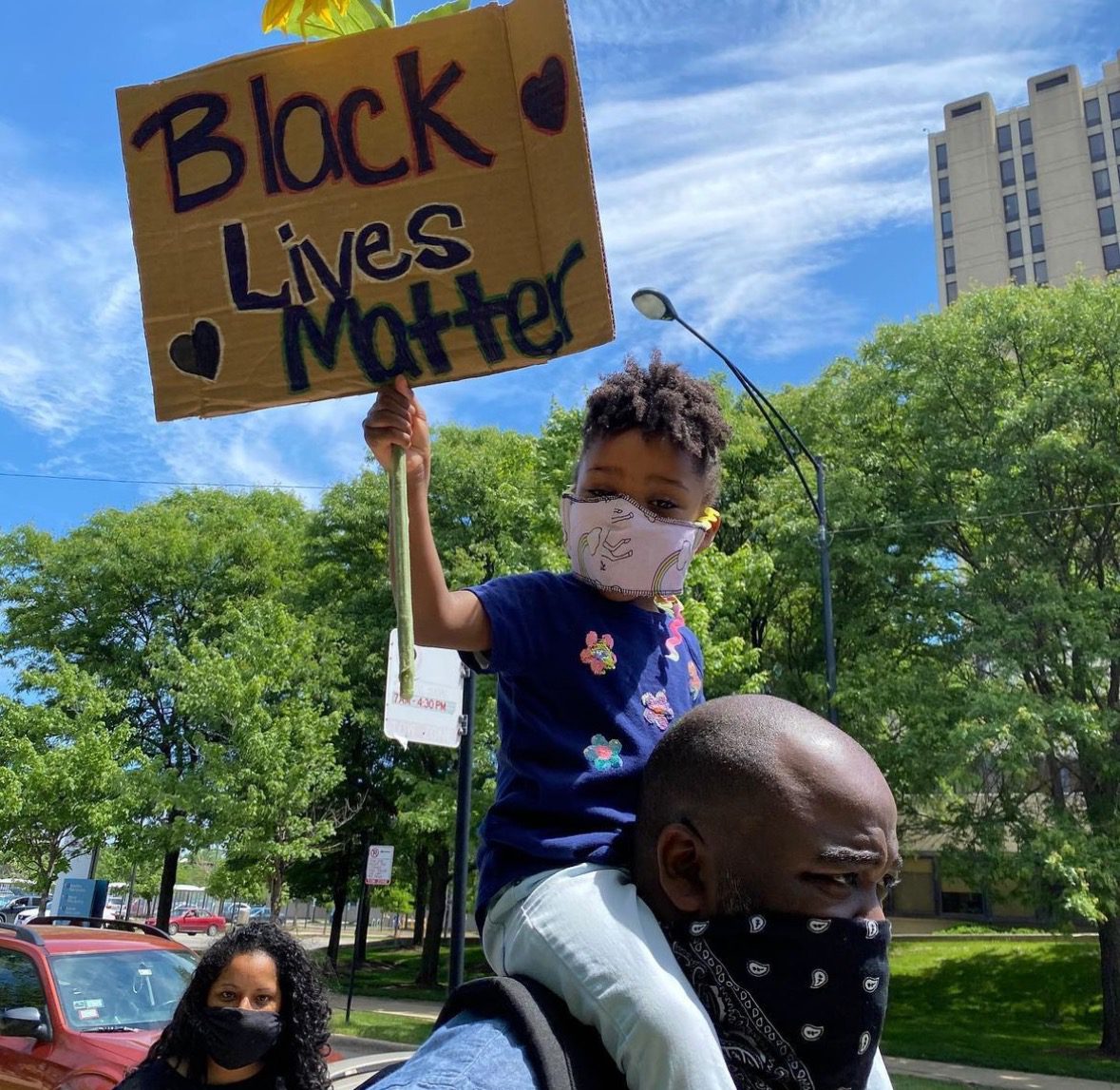 Photo shows a young child on Principal Jonas Cleaves' shoulders. They are in the middle of a protest outside and the child holds a sign that says "Black Lives Matter". Cleaves wears a black bandana over his mouth and the child has a pink mask on.