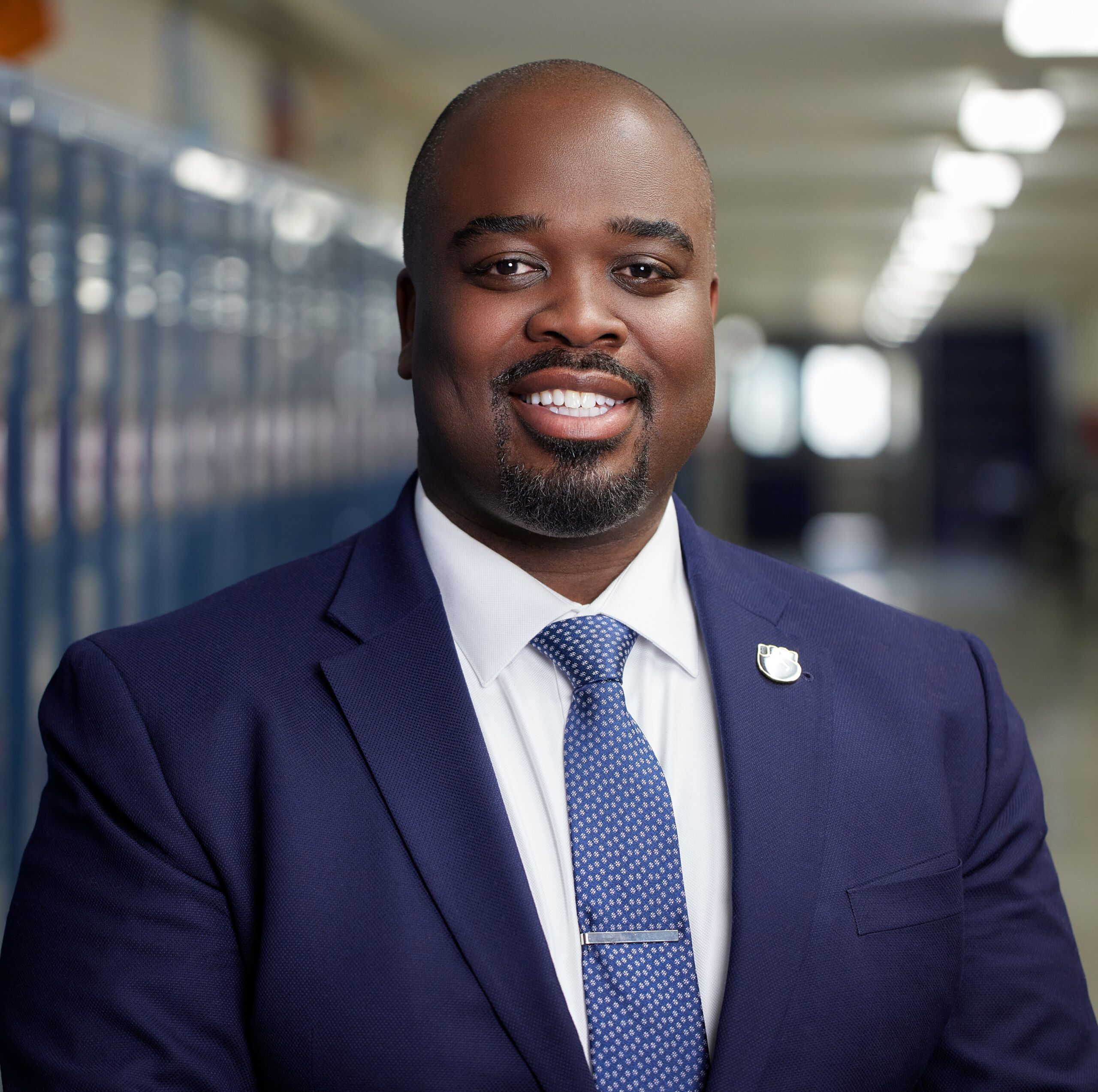 Photo is a professional headshot of Jonas Cleaves, principal of Johnson College Prep, in his school hallways. He is a Black man who is bald and is wearing a white button up shirt with a dark blue jacket and matching tie. He is smiling.
