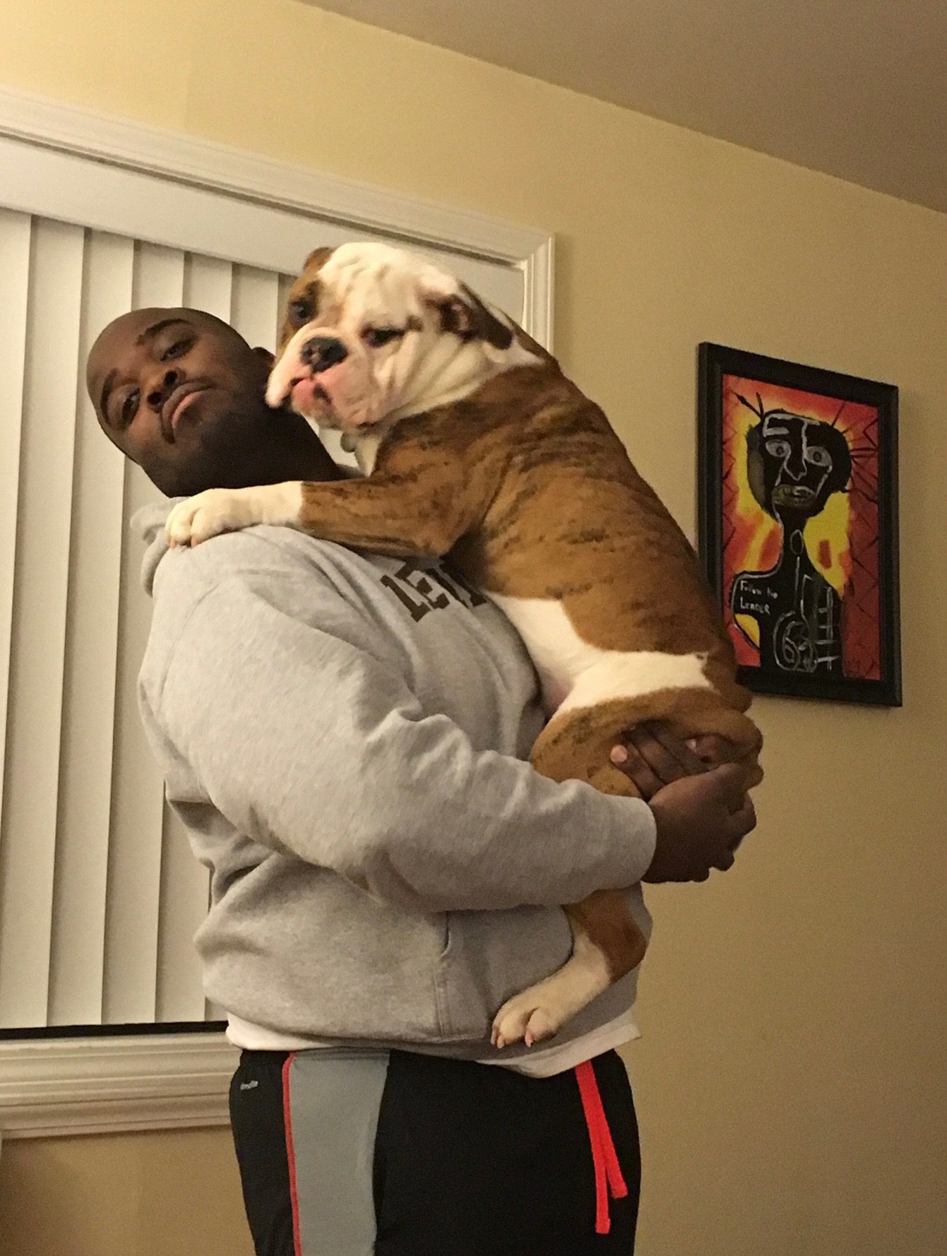 Photo shows Principal Jonas Cleaves standing and holding his big bulldog on his chest. The dog is hugging him. He is inside a house with a piece of art on the wall behind him.
