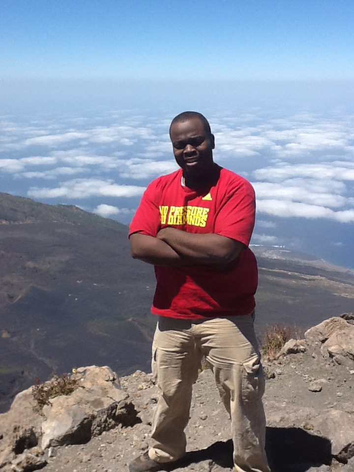 Photo shows Principal Jonas Cleaves standing on a mountain summit. You can see other mountains and clouds behind him. He is crossing his arms and standing with his legs apart -- a nice power pose.