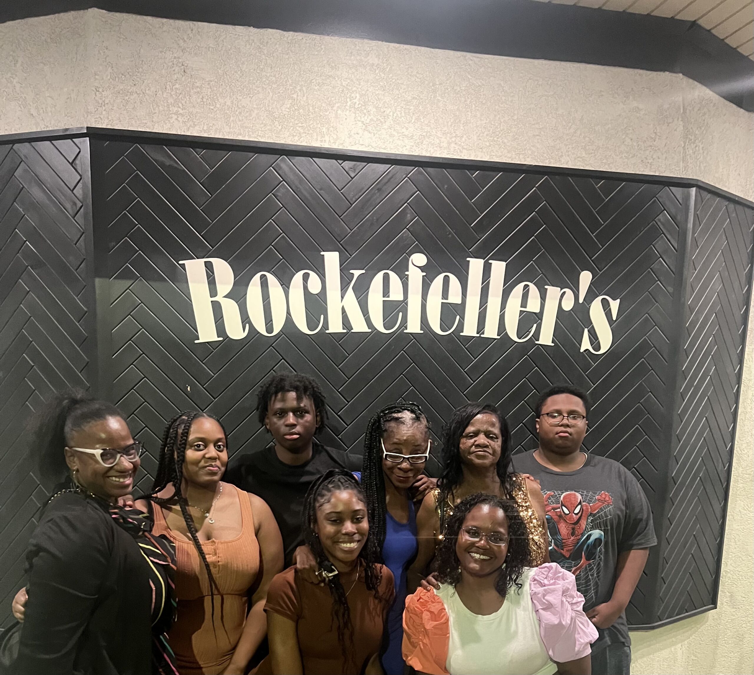 Photo shows Principal Kashawndra Wilson posing with her family in front of a black sign that has the word "Rockefeller's" on it in a bold white newspaper-like font.