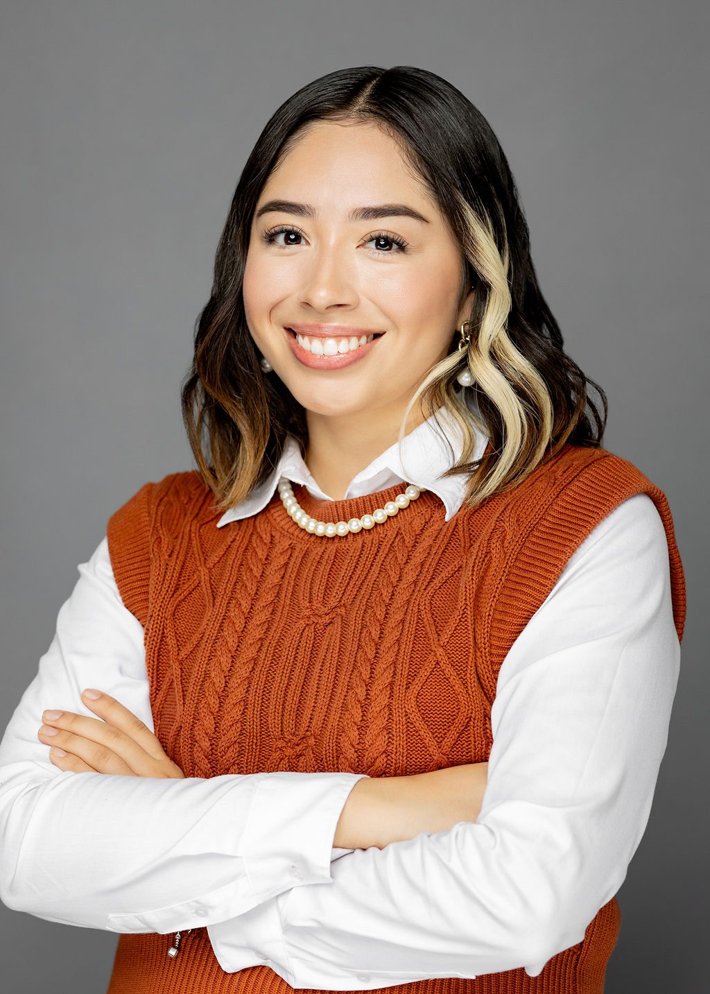 This photo is a professional headshot of Marlene Rios. She is a Latine woman with medium-length wavy dark brown hair with a few locks of blonde in the front. She is wearing a white long-sleeved button-up with a burnt orange sweater vest over it. She is also wearing a string of pearls around her neck and gold dangling earrings with big pearls at the end. She is smiling and crossing her arms confidently.
