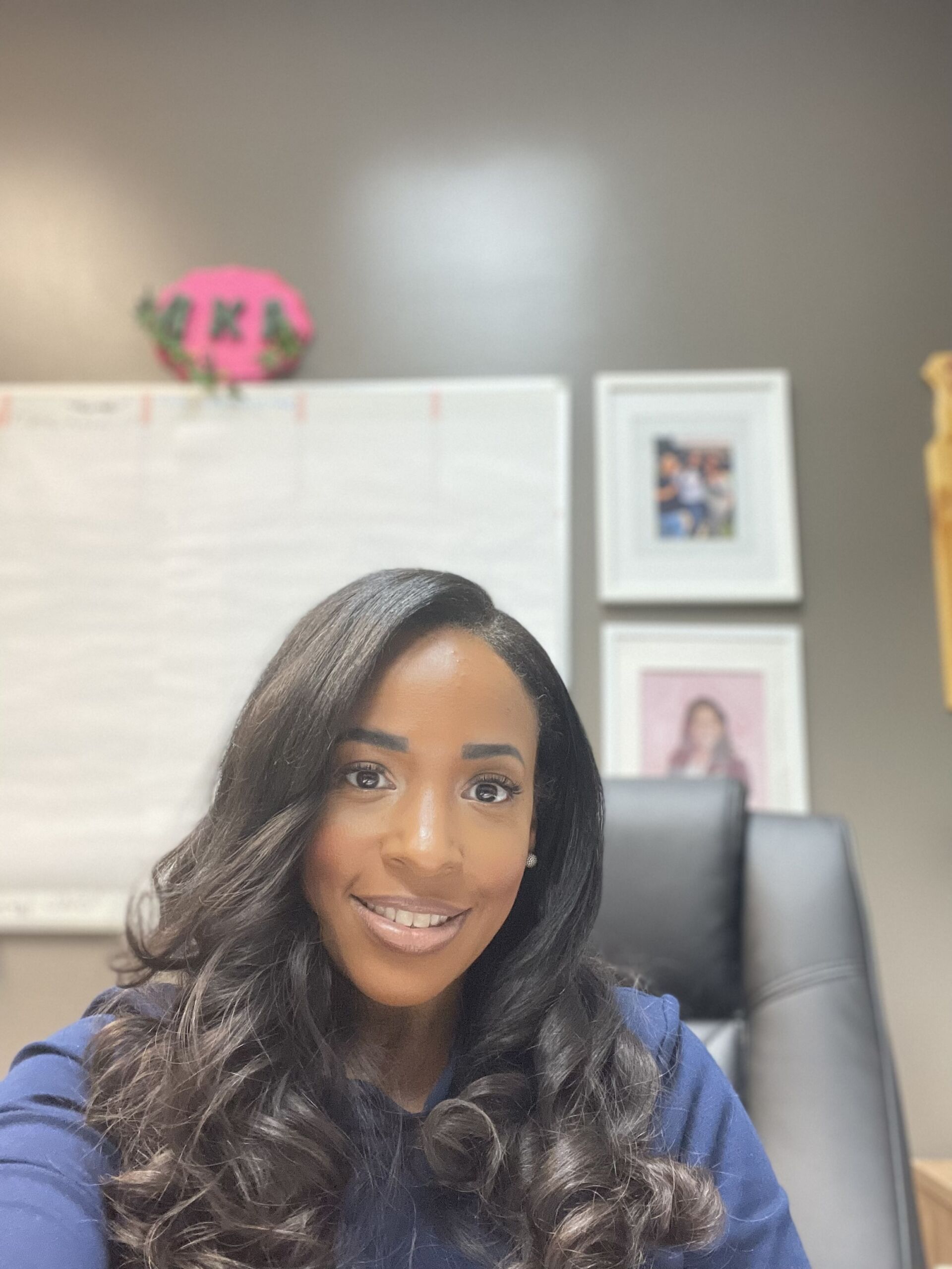 Photo shows a selfie of Principal Miracle Moss in her office at Rowe-Clark Math and Science Academy. She is smiling at the camera as she sits in her office chair. Her hair is in loose curls and she is wearing a dark blue blouse.