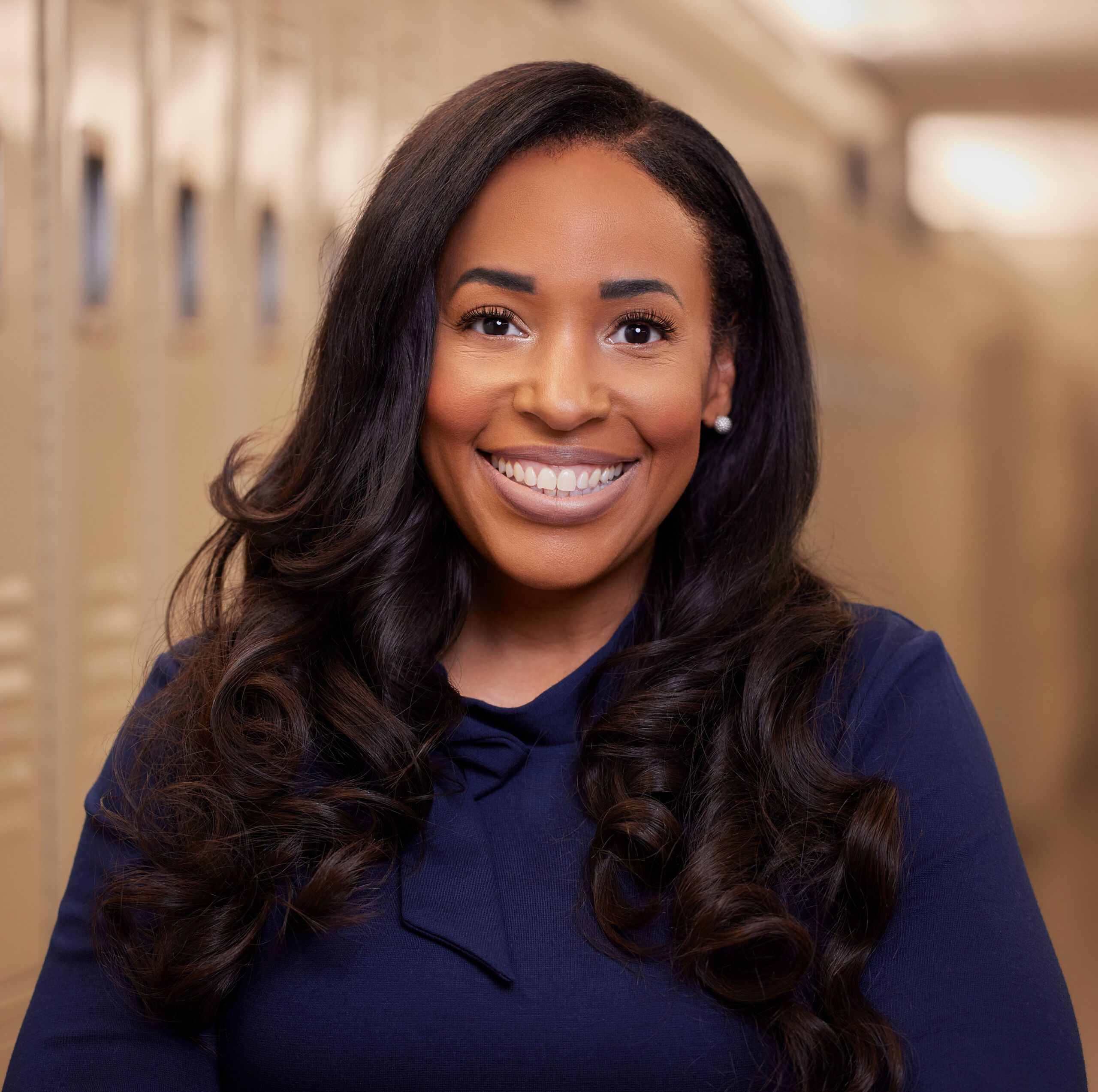 Photo shows a professional headshot of Miracle Moss, principal of Rowe-Clark Math and Science Academy. She is a Black woman with long straight black hair. She is smiling and wearing a dark blue long-sleeved blouse with a bow on the neckline. In the background, you can see lockers in a hallway of Rowe-Clark.