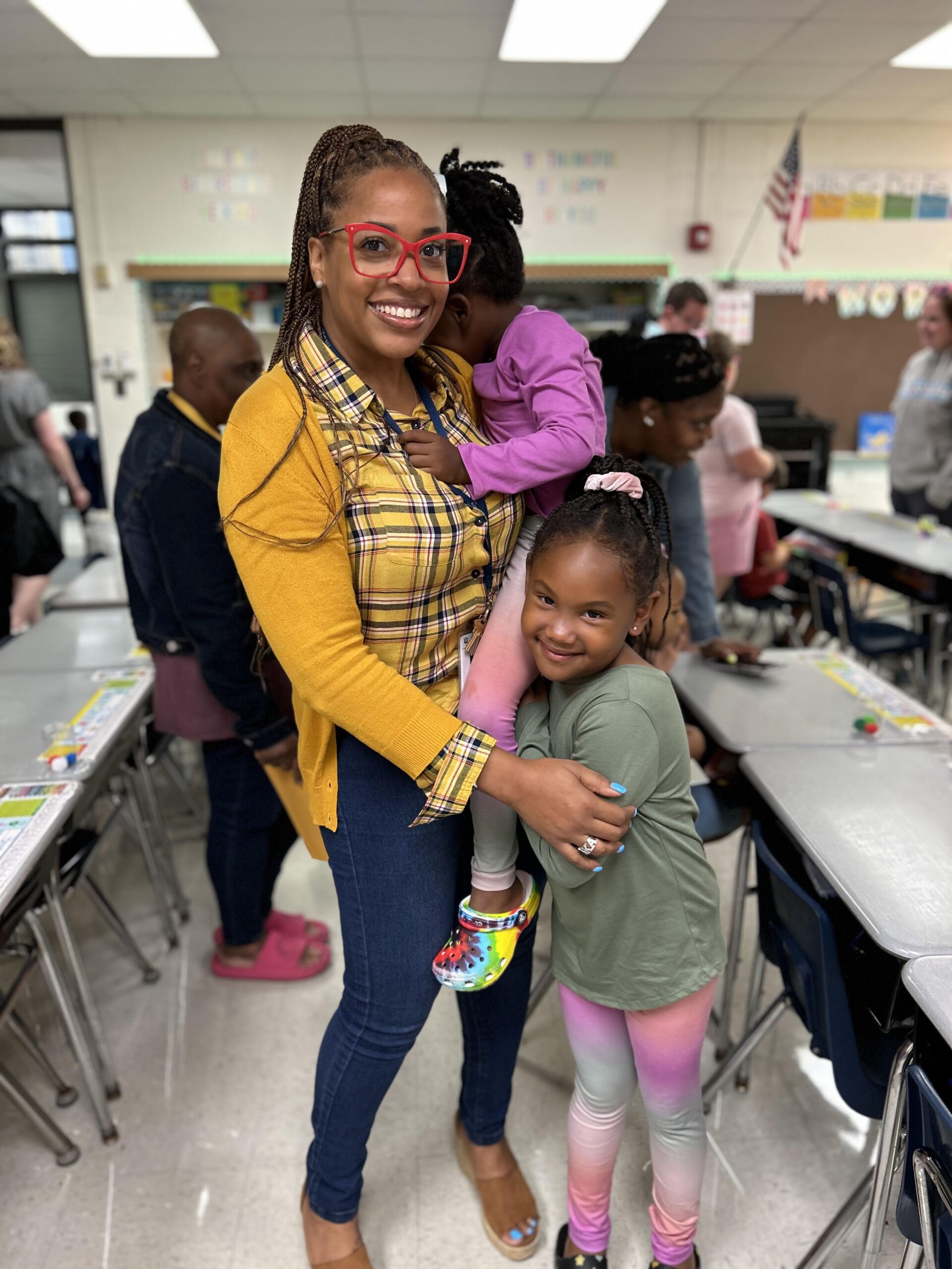Photo shows Principal Miracle Moss with her two kids in a school classroom. She is holding her youngest child, who is hiding her face from the camera. Her other child is next to her leg, side-hugging her and smiling at the camera. Moss is also smiling at the camera. In the background, you can see elementary school desks and other adults and children moving through the space.