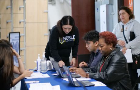 Image shows a Noble School parent and a student/alum sitting at a table and working on laptops to fill out the Chicago Public Schools family survey for Noble's charter renewal. Noble's financial Controller, Mireya Diaz, stands next to them, helping them with something on the computer. Another parent stands in the background, watching.