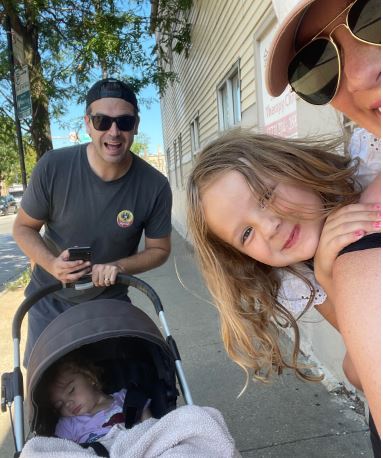 Photo shows a selfie of Principal Nora Lawrence and her family as they are outside on a walk. One of her daughters is on her back and the other one is in a carriage that her husband is pushing.