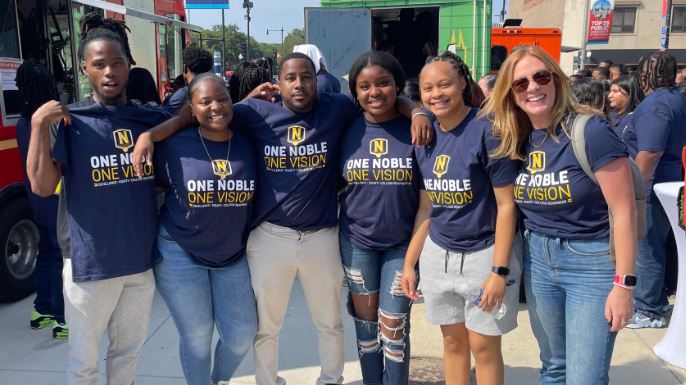 Photo shows Principal Nora Lawrence posing with Noble alumni that she taught who have all come back as staff members. They are all wearing Noble shirts, having just come out of the Noble Schools' Staff Kickoff event for the 2023-2024 school year.