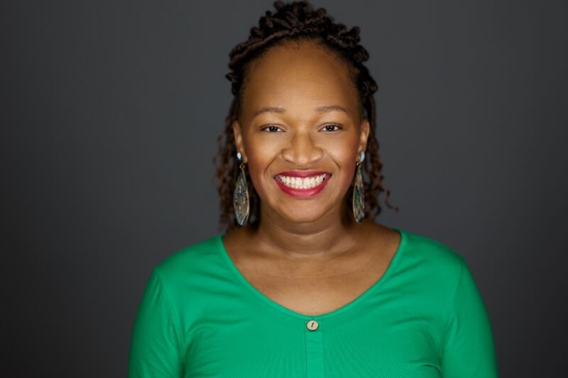 Professional photo of Pinkston, a black woman with twists and curls in her hair. She is wearing a green top and smiling.