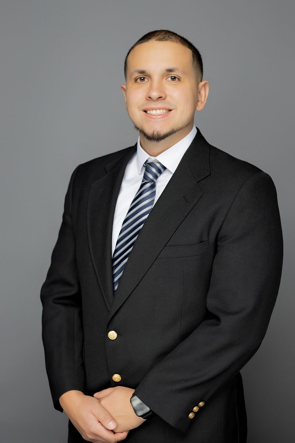 This photo is a professional headshot of Ricardo Quezada. He is a Latine man with a very short black buzzcut and beard. He is wearing a white button-up with a black blazer buttoned on top of it. He has on a black and silver striped tie. He is also wearing a black smartwatch on his wrist. He is smiling and has his arms relaxed at his sides with his hands clasped in front of his waist.