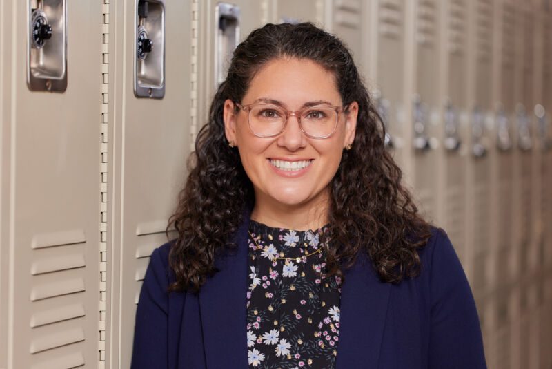Photo is a professional headshot of Carrie Spitz, principal of Pritzker College Prep, in her school hallways. She is q Latina woman with curly hair and is wearing a black floral top with a black jacket. She is smiling.