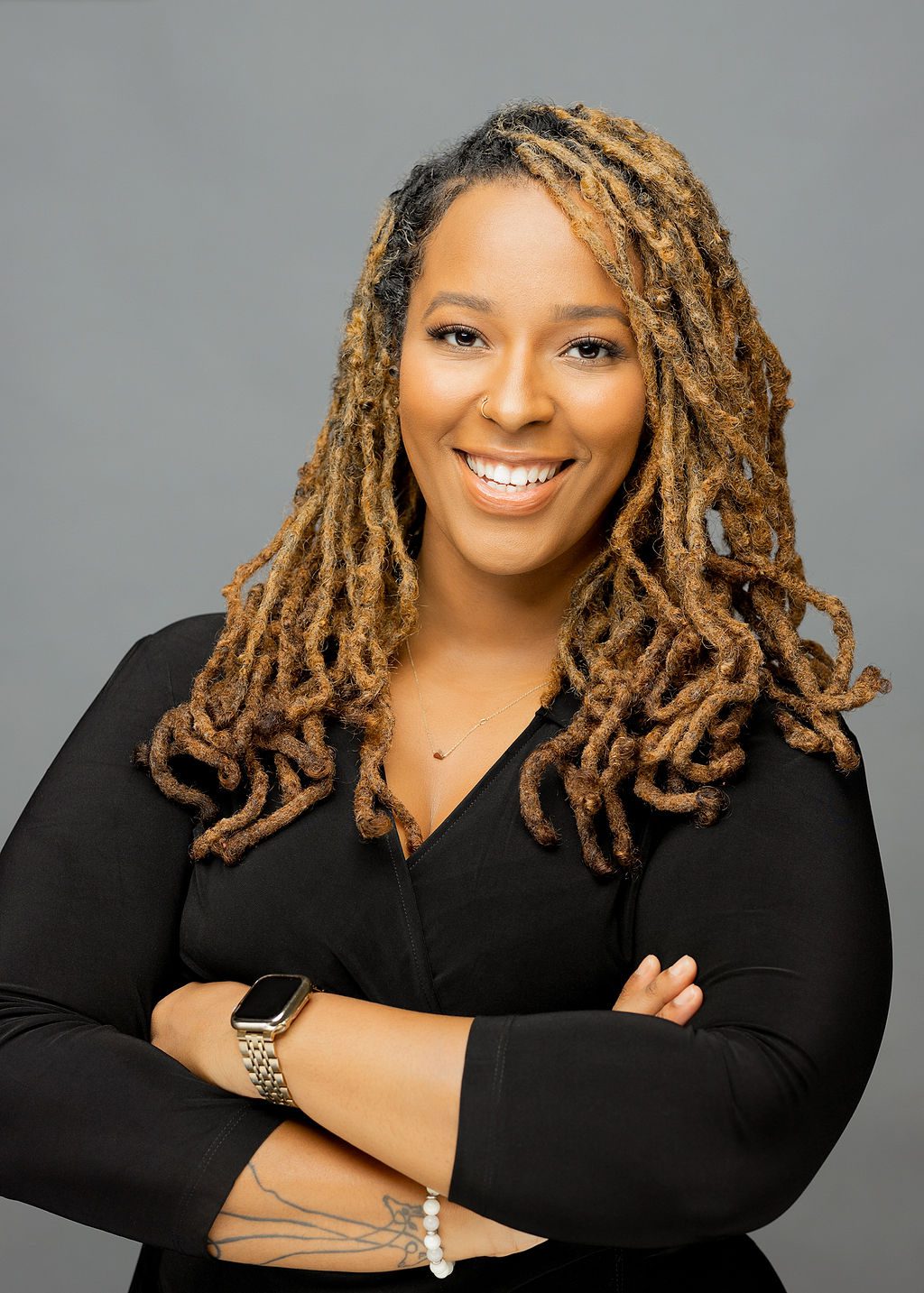 This photo is a professional headshot of Tyesha Thomas. She is a Black woman with long, curly light brown locs. She is wearing a medium-sleeved black blouse, a thin silver chain with a small red gem at the end, a gold smartwatch on one wrist, and a white beaded bracelet on the other. She is smiling with her arms crossed confidently.