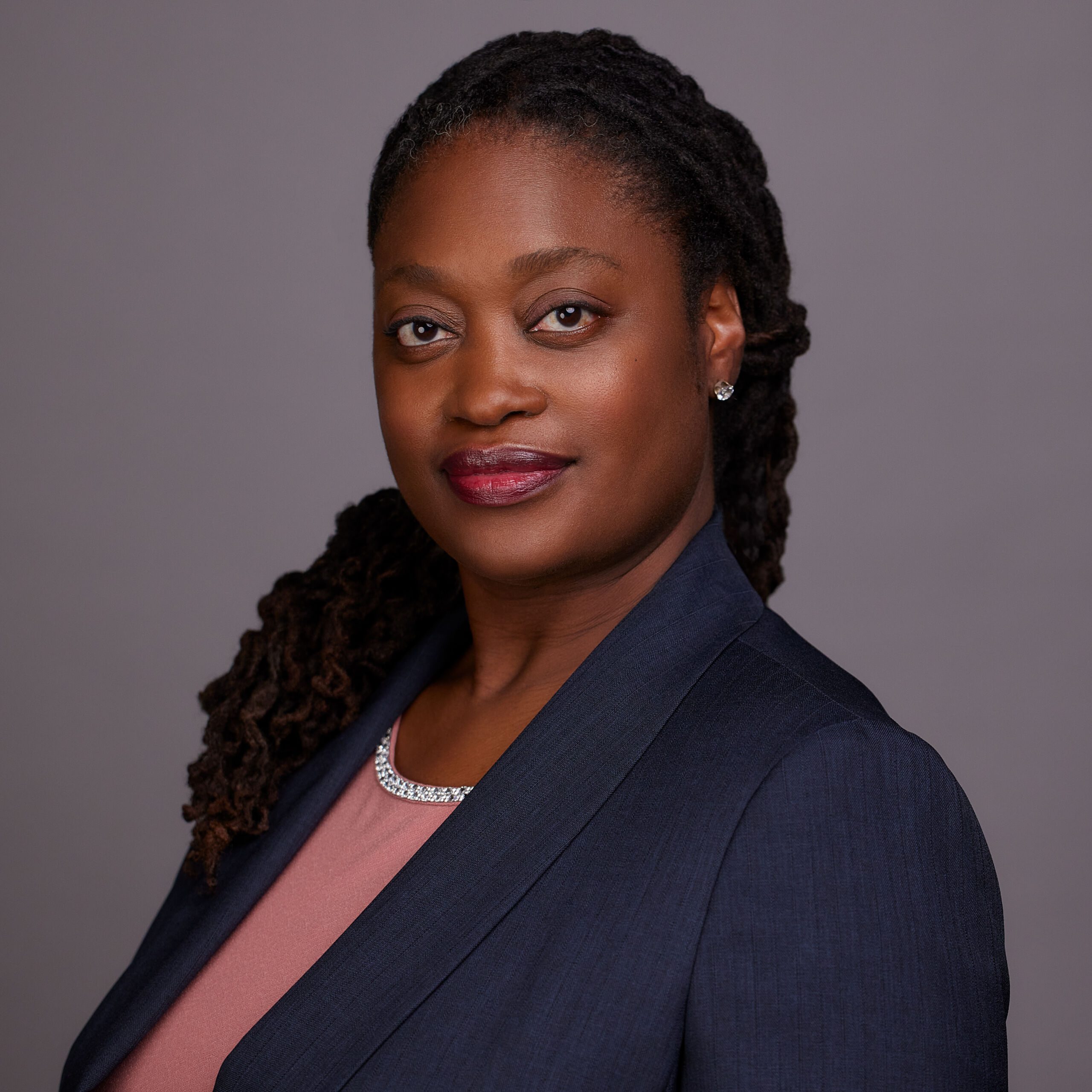 Photo shows a professional headshot of Tina Ellis, principal of DRW College Prep. She is a Black woman with long black hair. She is wearing a pink blouse with a navy blazer. There is a blank gray background behind her.
