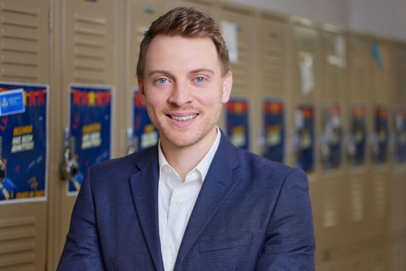 Photo is a professional headshot of Ben Gunty, principal of Noble Street College Prep, in his school hallways. He is a White man with short brown hair and is wearing a white button up shirt with a dark blue jacket He is smiling.