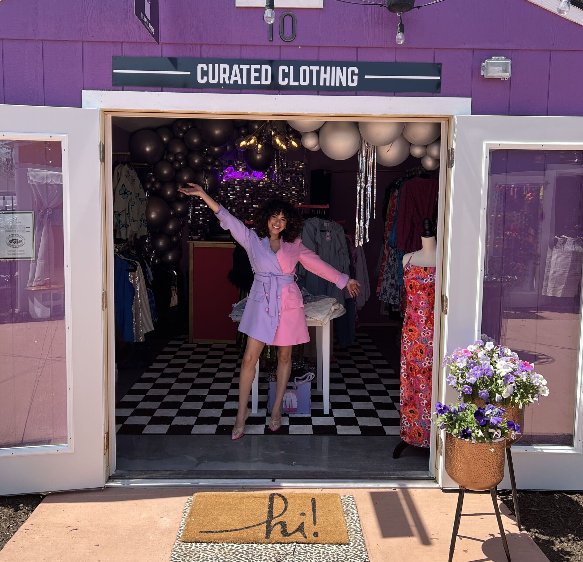Photo shows Karina Estrada, a Noble Schools alum and owner of Dear Me Fashion Boutique, standing with her arms outstretched in a celebratory manner in the middle of her temporary shop location for Dear Me at Berwyn Shops. The shop is in a small purple building and the double doors are wide open. A welcome mat is in front that says "hi!". Karina is wearing a short dress with long sleeves that is cut in half by color: lavender on the left and bright pink on the right. A black and white sign above the doors says "Curated Clothing"