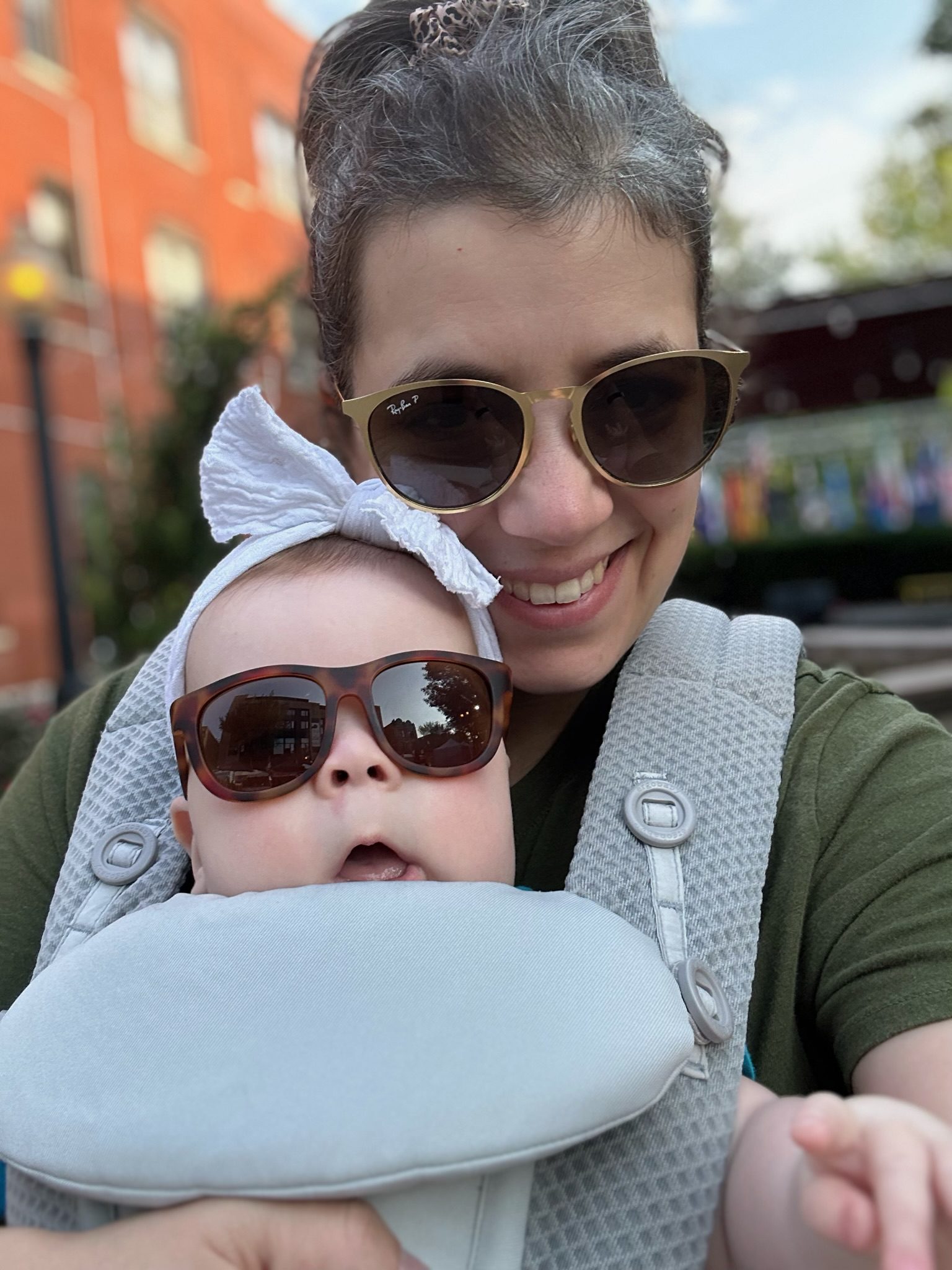 moya with a baby strapped to her chest smiling, both wearing sunglasses