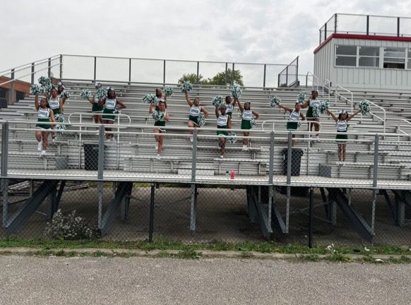 Cheerleaders in the stands shake their pom poms and cheer on the team. They are wearing green and white cheer tops and skirts.