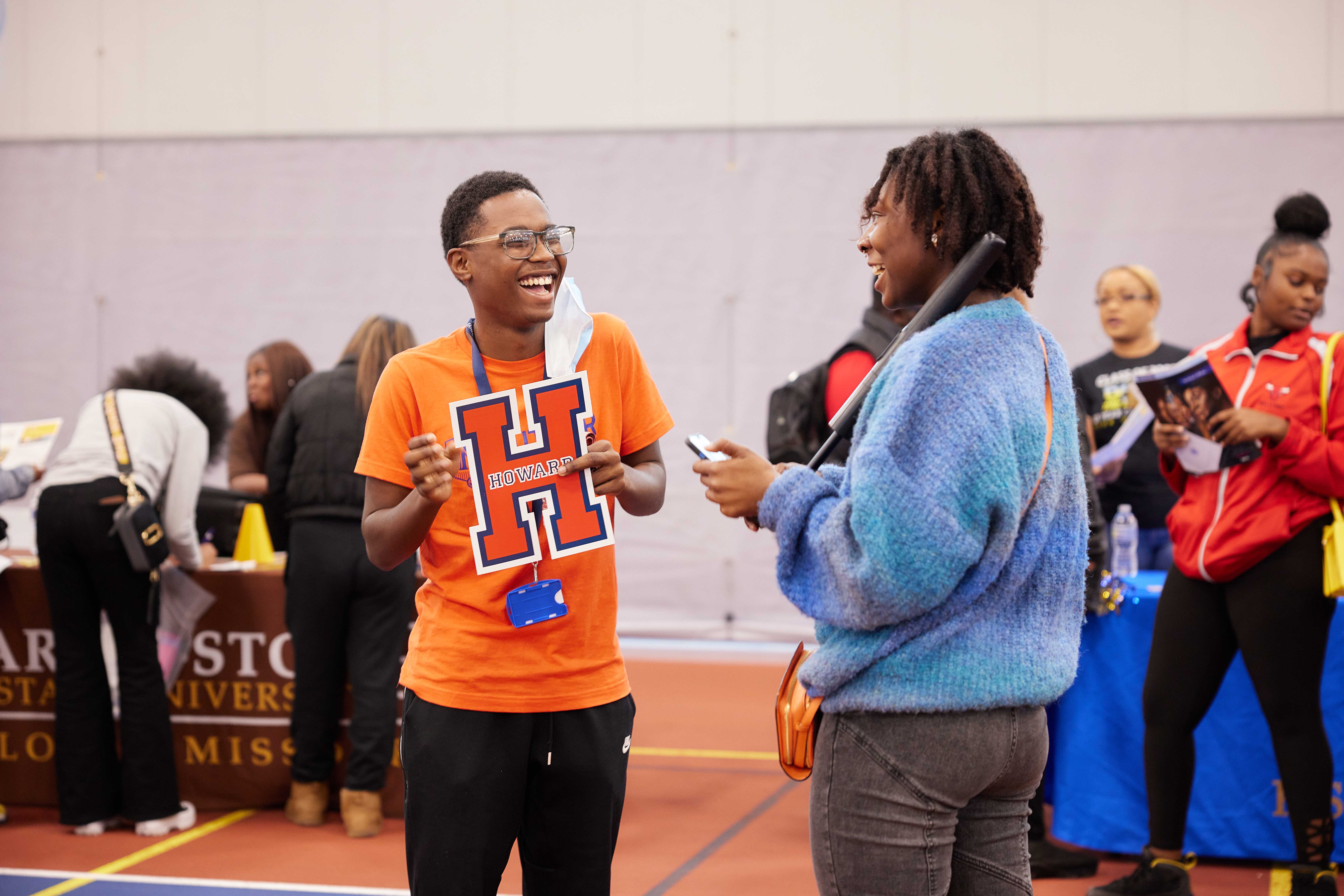 In this photo, you can see Antwan H, a senior at Butler College Prep and a young Black man, smiling and laughing as he talks with a Butler alum at the HBCU College Fair. He is holding a big Howard University decal in his hands. Behind him and the alum, you can see other students and college representatives at the fair.