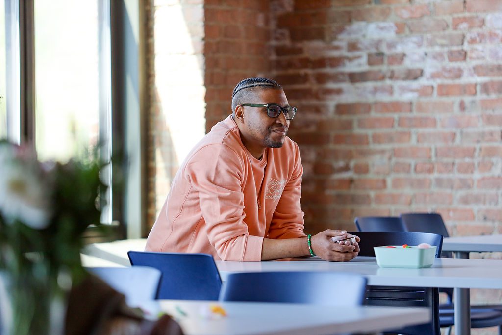 In this photo, D Nigel Green, Director of Equity, Inclusion and Diversity at Noble Schools, sits at a small table, smiling gently and looking to the right at something off camera. He is a Black man with his hair in short cornrows on the top and shaved on the sides. He is wearing glasses and a peach long-sleeved sweatshirt. He is watching the graduation of the 5th cohort of Noble Schools' Diverse Leaders Fellowship.