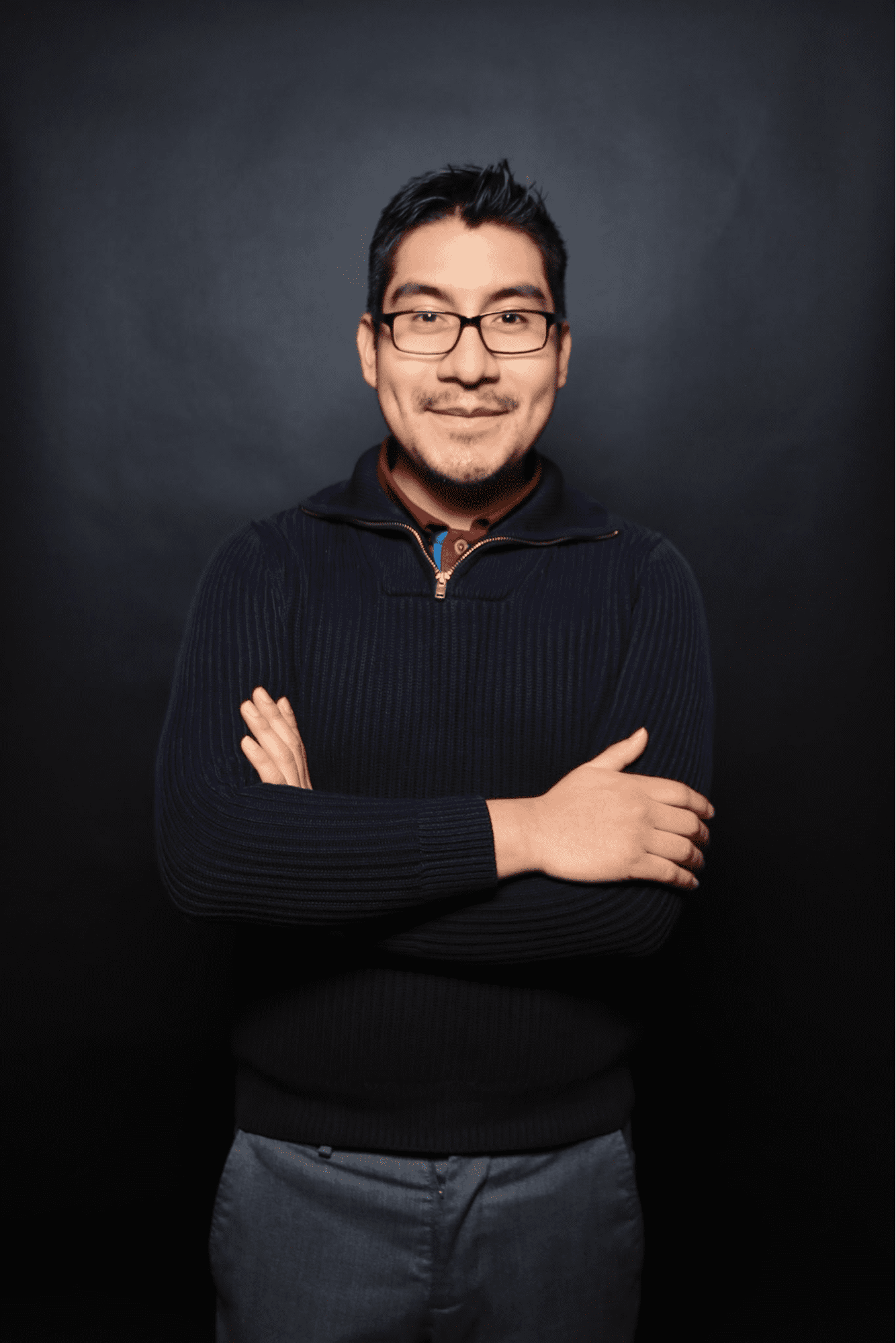 Professional photo of David Rios- Perez he has short dark hair and is wearing glasses. He has on a navy blue sweater with grey slacks. His arms are crossed.