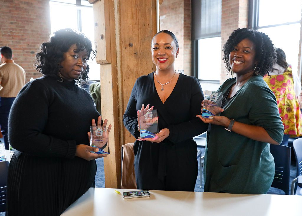 In this photo, you can see three women of color standing in a room, presenting crystal plaques in their hands. They are smiling at and posing for the camera. On the left in Tanisha Hall, a teacher at ITW David Speer Academy. She is a Black woman with curly mid-length black hair. She is wearing a long-sleeved black sweater dress and big earrings. To her right are two other members of the 5th cohort of Noble Schools' Diverse Leaders Fellowship—Joy Pryor, a Latine woman, and Dr. Alyssa Hamler, a Black woman. They are both also wearing formal wear. Behind them all, you can see a bright sunny room where other people are mingling in groups.