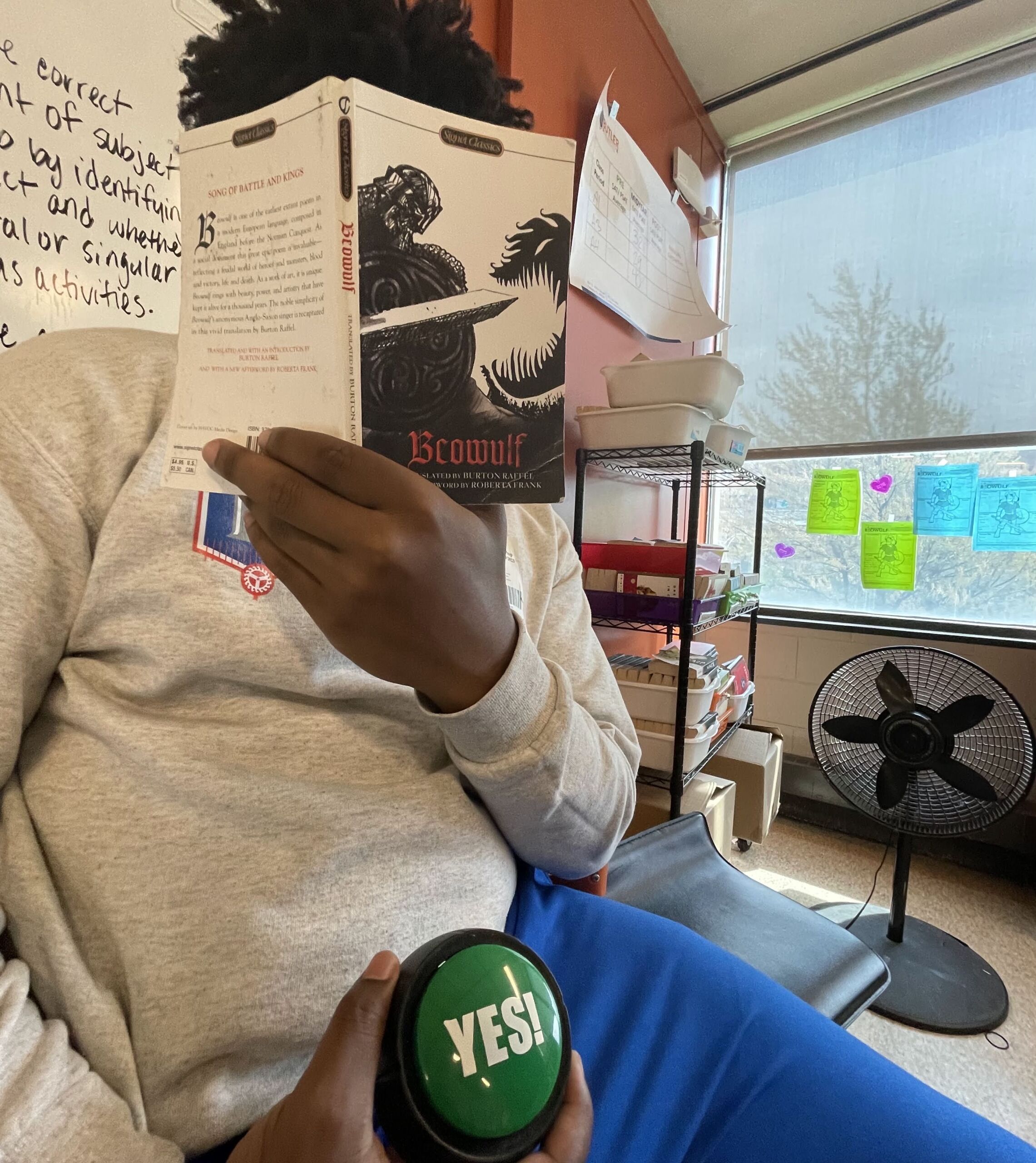 In this photo, you can see a Black student with a Butler College Prep sweatshirt on reading a book titled Beowulf. They are holding the book in front of their face with one hand and a green button in their other hand that says "Yes!". Behind them, you can a classroom space with a whiteboard, some shelves, and a window.