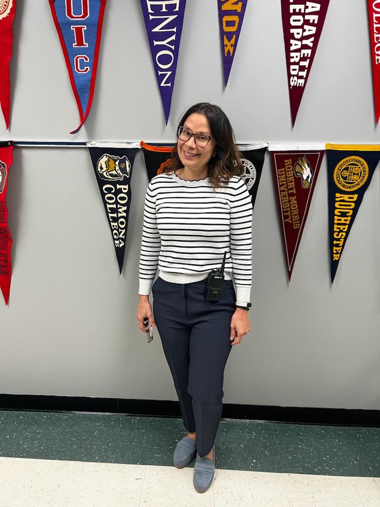 In this photo, you can see Ms. Montero, a Latina woman, standing in the hallway at Noble Street College Prep. She is standing in front of some college pendants that are attached to the wall. She is smiling and wearing glasses, a black and white striped sweater and dark blue slacks. A walkie talkie is attached to her hip.