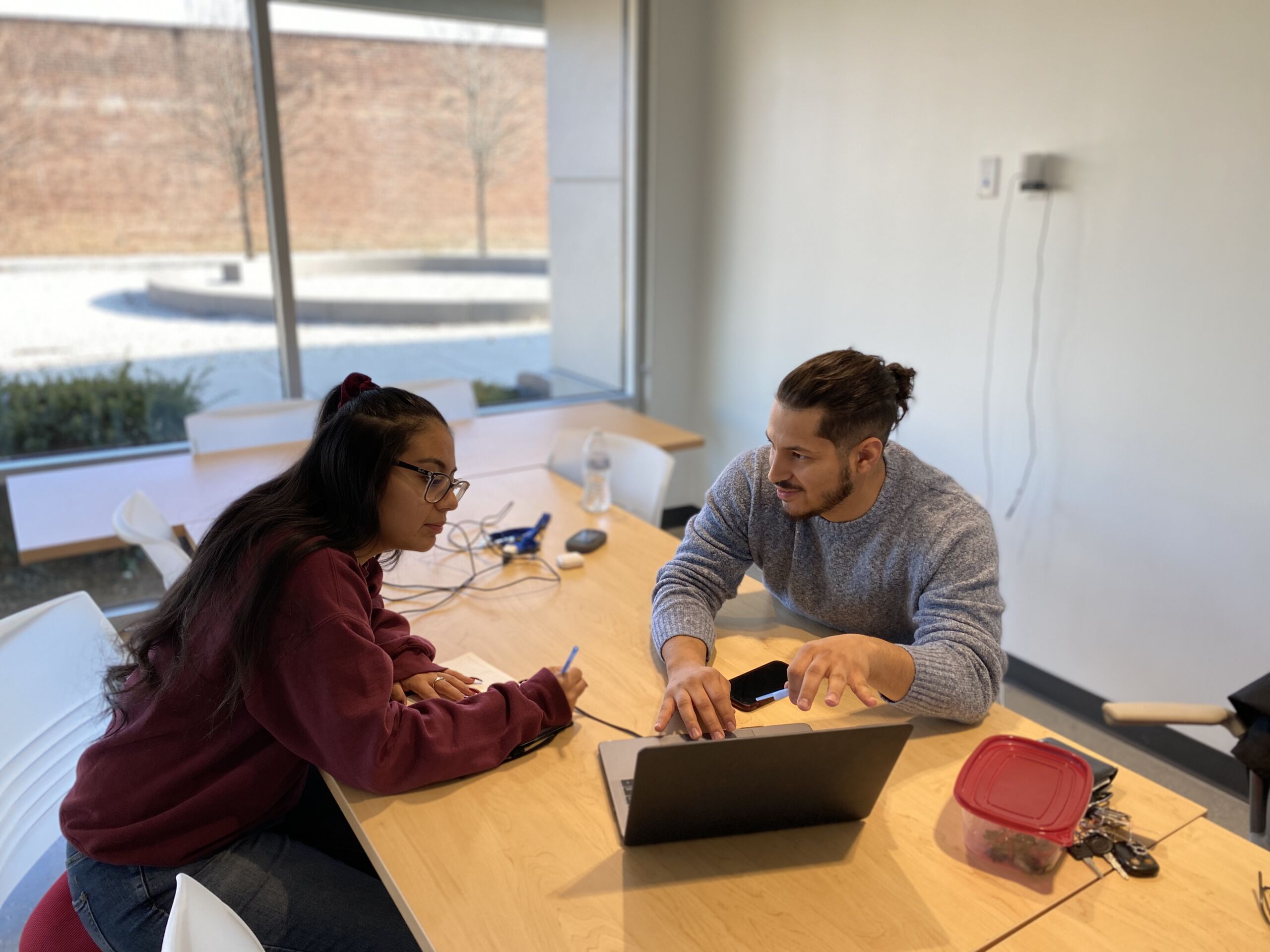 In this photo, you can see Isaiah Martinez sitting at a table with a young Noble Schools' alum, walking her through something on a laptop. He is looking at her as he points at something on the laptop. She has her head leaned forward as she looks at the laptop and takes notes with pen and paper.