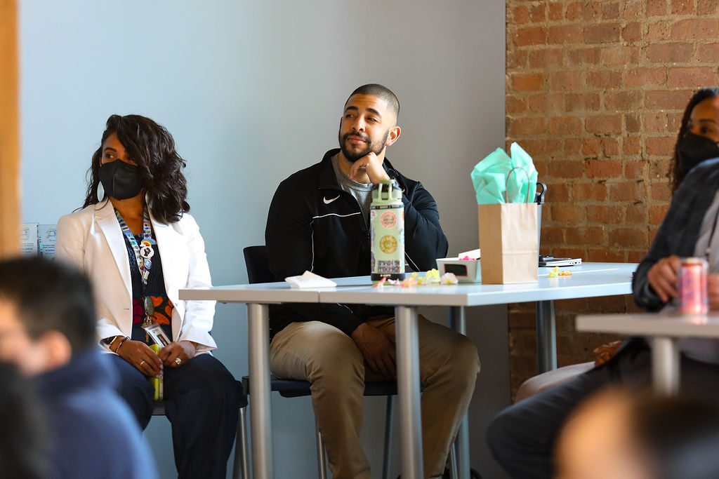 In this photo, you can see Jacob Goldstein, assistant principal of culture at Chicago Bulls College Prep, sitting at a table with other people and looking to the left at something off camera. He is a Black man with a beard and black hair that is closely shaved. He is wearing a black Nike athletic jacket and khakis. He is smiling slightly in this photo as he watches the Diverse Leaders Fellowship cohort 5 graduation.
