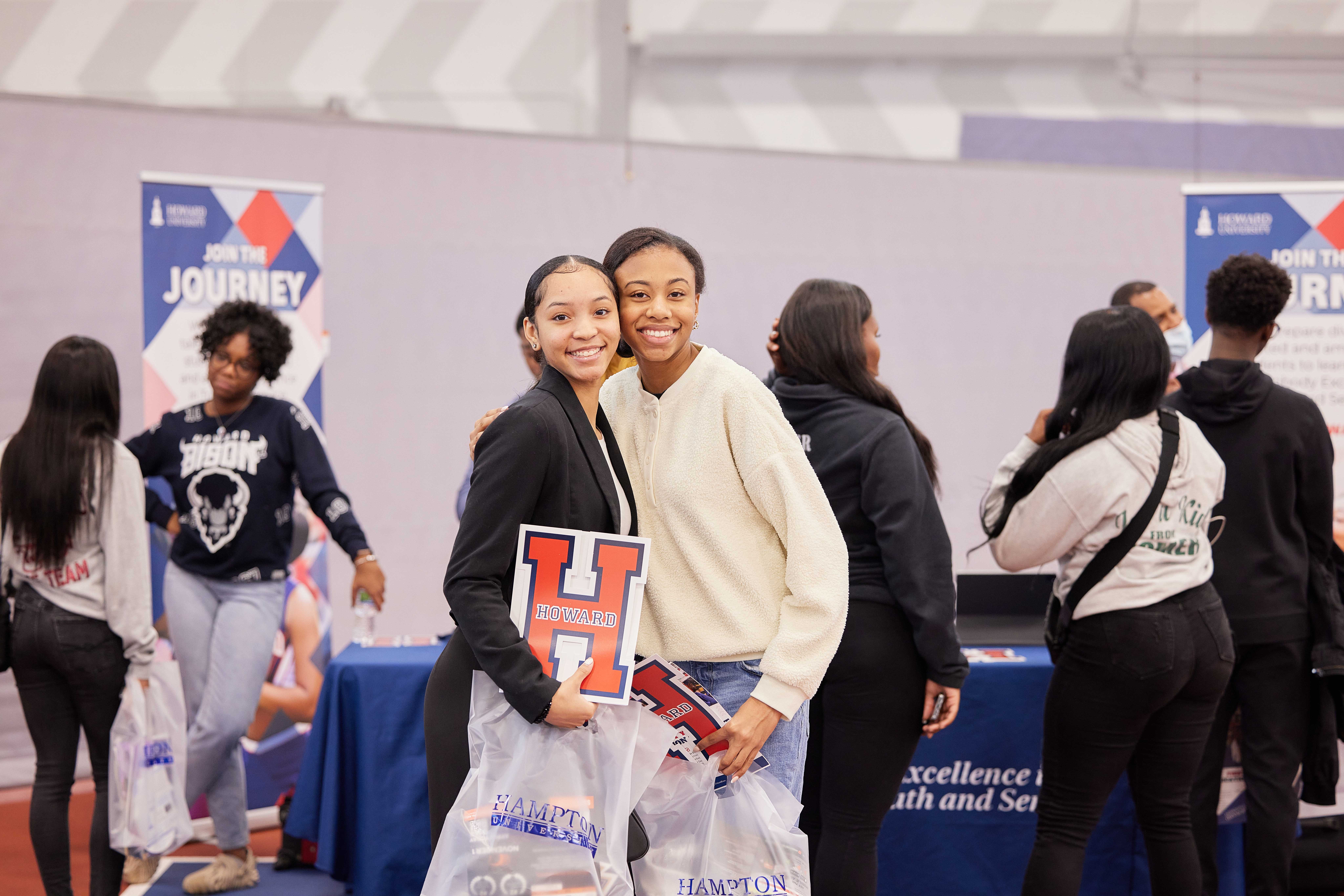 In this photo, you can see Madison and Laniya, two seniors at Muchin College Prep, side-hugging and smiling as they pose in front of the Howard University booth at Noble Schools' HBCU College Fair. Madison is holding a big decal for Howard University right in front of her waist prominently. Laniya also is holding one but it is partially hidden. They both are holding goodie bags that have Hampton University's logo on it. Behind them, you can see Howard University college representatives talking with other Noble students.