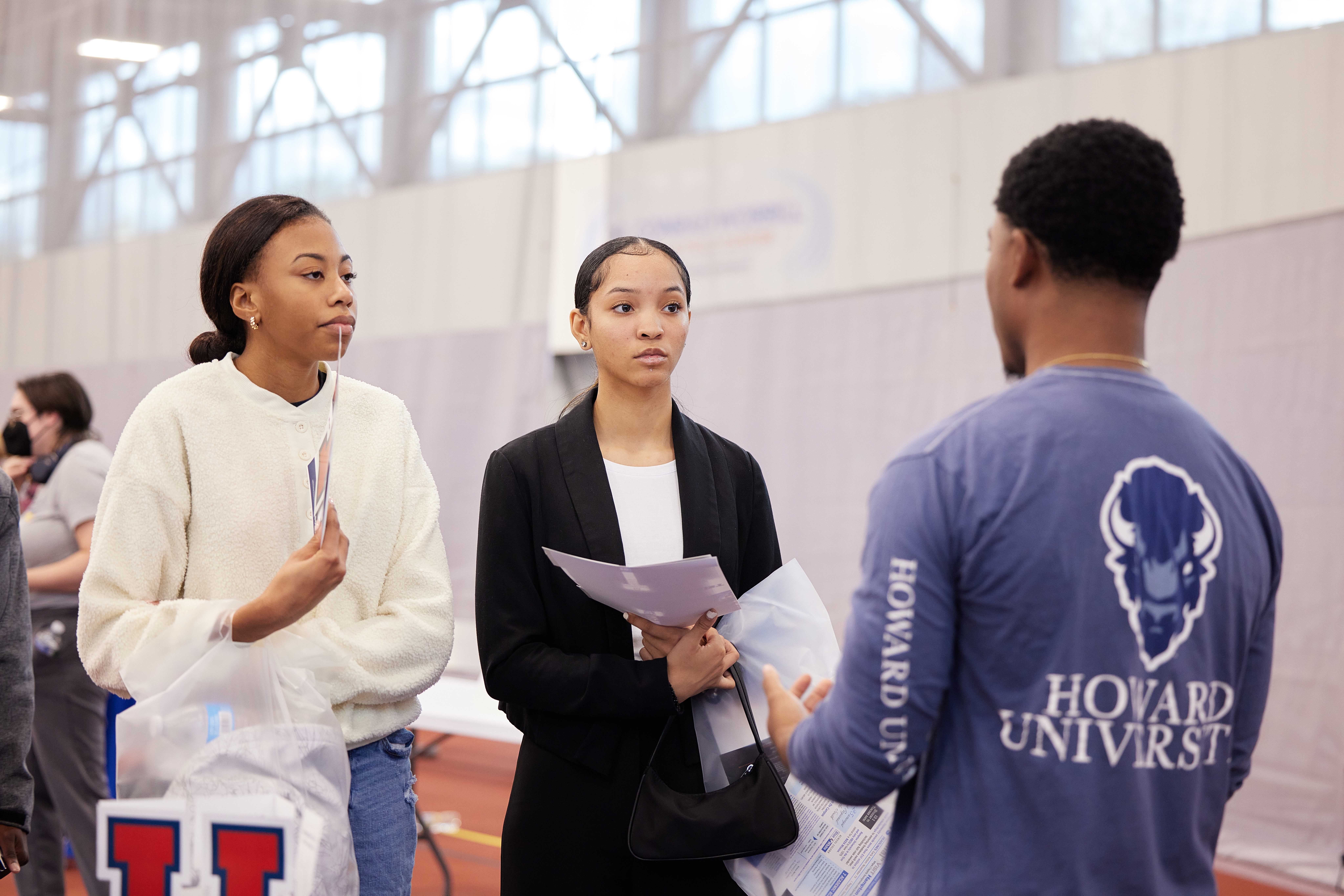 In this photo, you can see Laniya and Madison listening intently to a Howard University college representative. Both of them are young Black women and are dressed nicely. Madison has on a black suit with a white blouse and Laniya is wearing a nice white sweater with jeans. They both are holding various bags and printed materials from different universities and colleges at the fair.