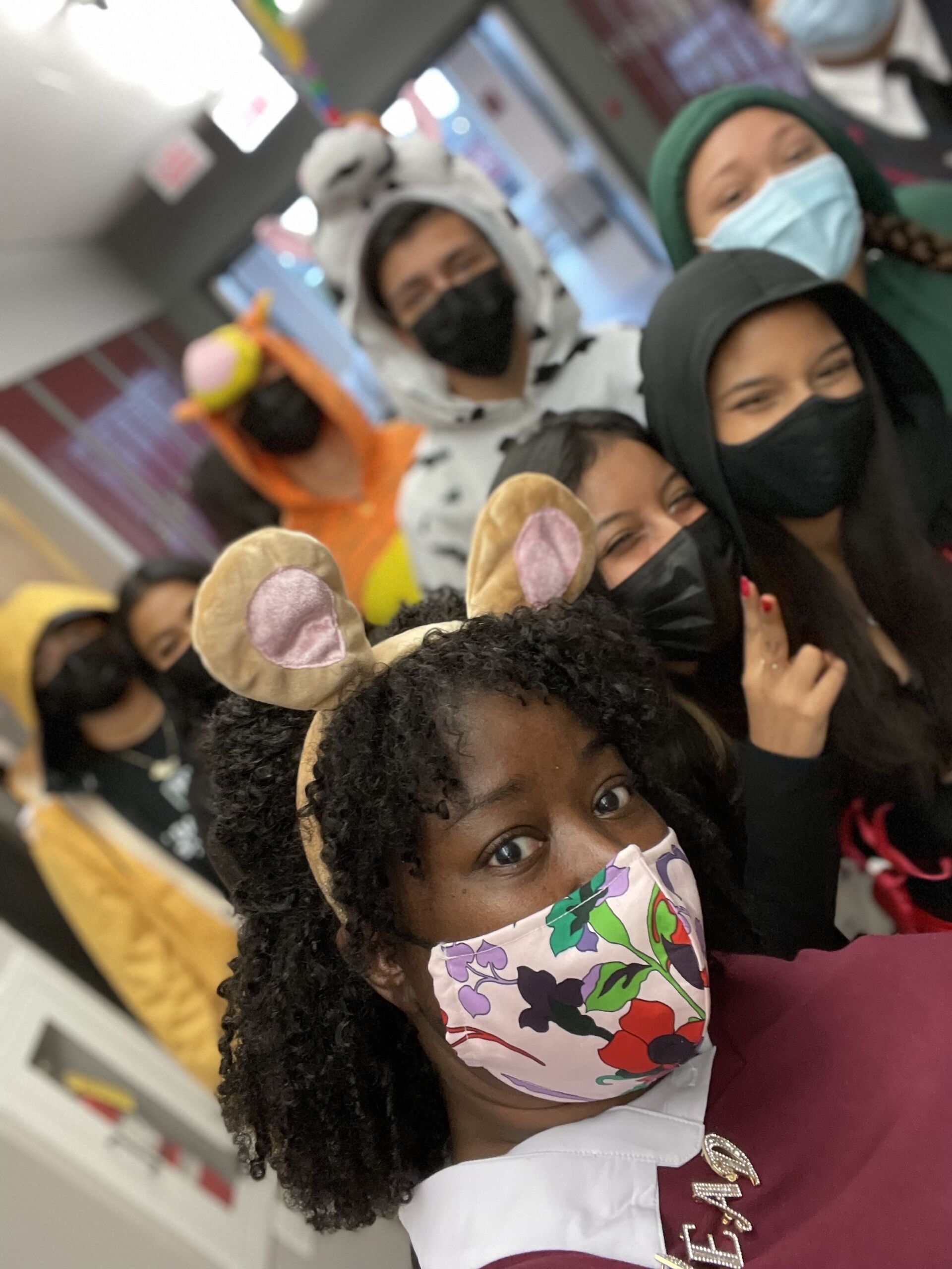 In this photo, you can see Tanisha Hall dressed up as Ms. Read from Arthur with some of her Speer students, some of them also dressed up in costumes, standing behind her. They are all posing for a selfie together in the school hallway.