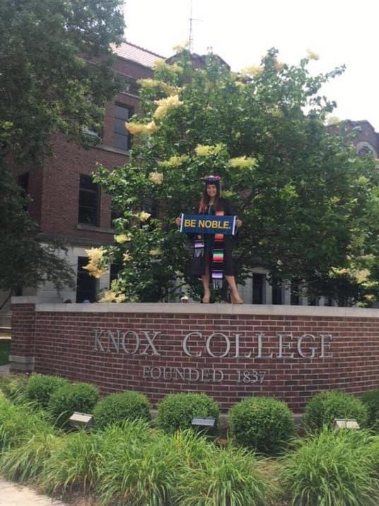 In this photo, you can see Sbeydi Gonzalez standing on top of her college's campus entrance sign—which reads "Knox College, Founded in 1837". She is wearing her graduation robes and cap and is holding a little handheld sign that reads "Be Noble".