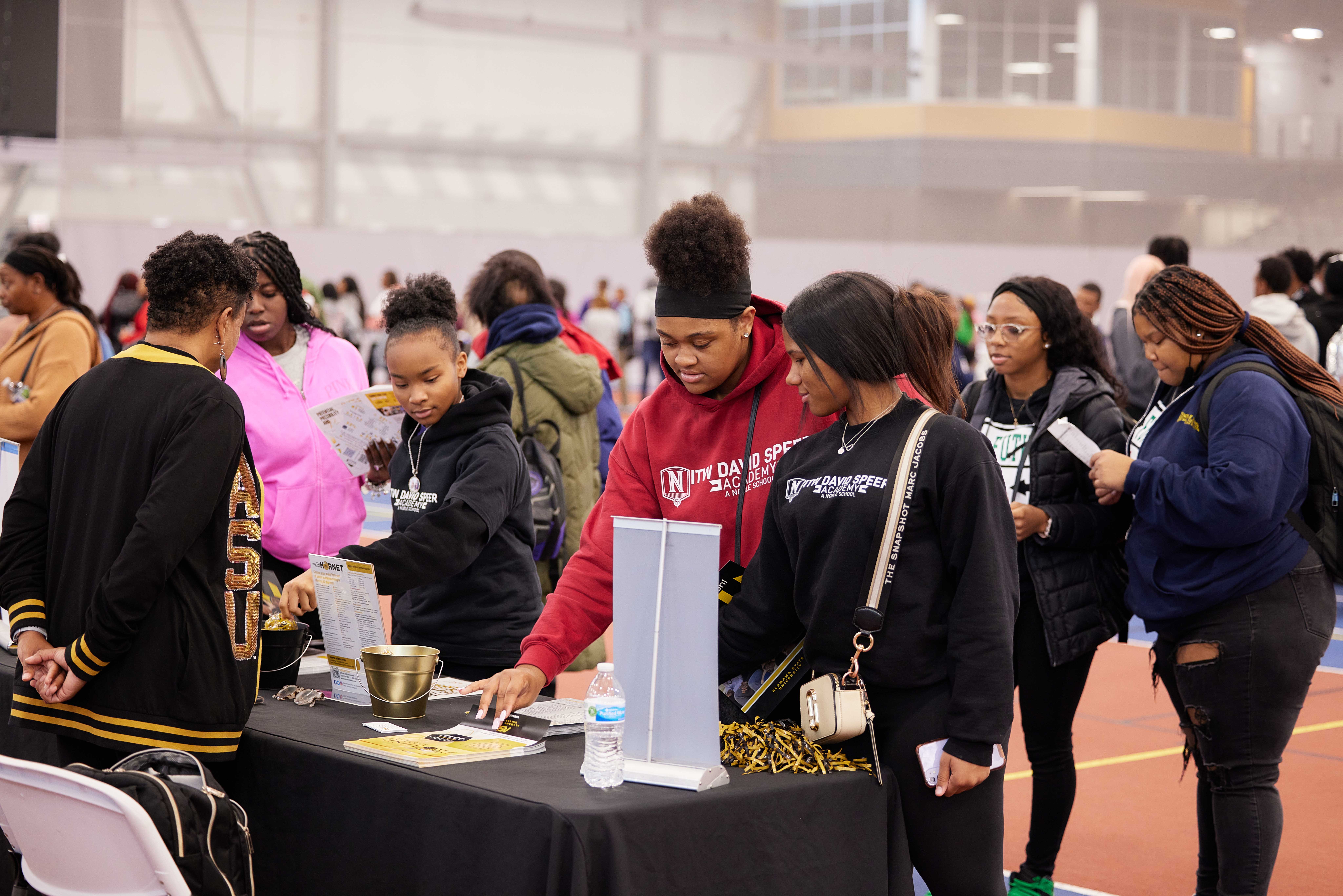In this photo, you can see a handful of Noble Schools' students from different campuses looking at and grabbing printed flyers and materials at a college recruitment booth for Alabama State University, an HBCU. Behind them, you can see several other students wandering around a large indoor track field, visiting other HBCU college booths. The students most in the foreground of this photo are wearing ITW David Speer Academy sweatshirts.