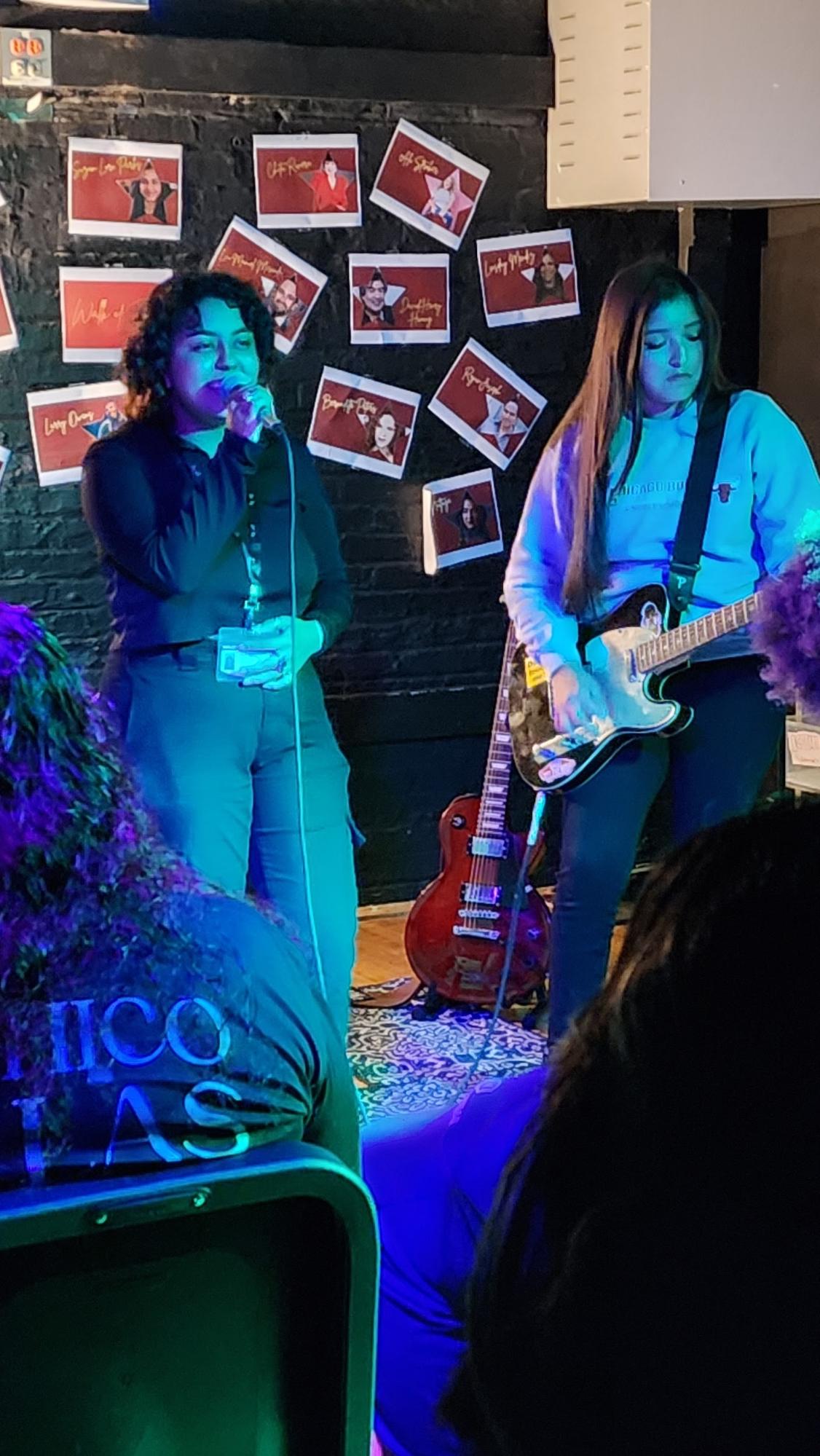Students performing at open mic night. One is singing and wearing dark-colored clothing and the other is playing the guitar. A blue light is shining on both students.