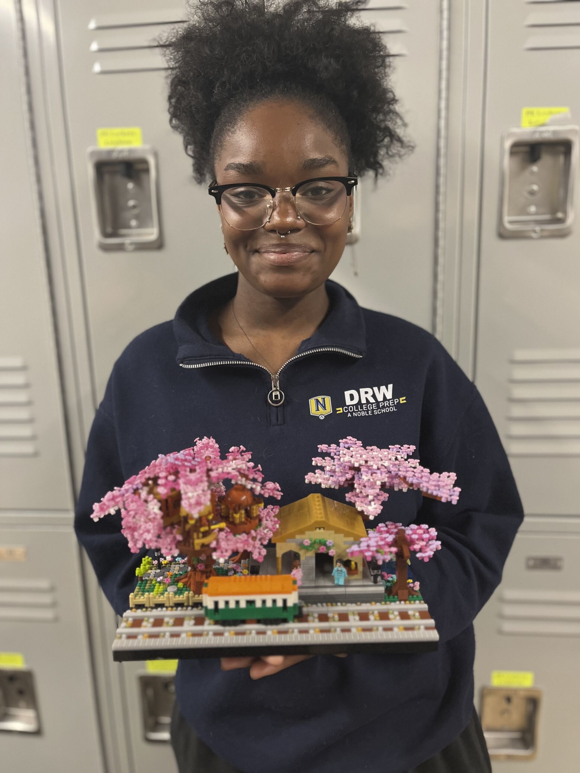 DRW student holding a house and surrounding front yard with pink trees, all made of Legos. the student is wearing a navy sweater standing in front of lockers