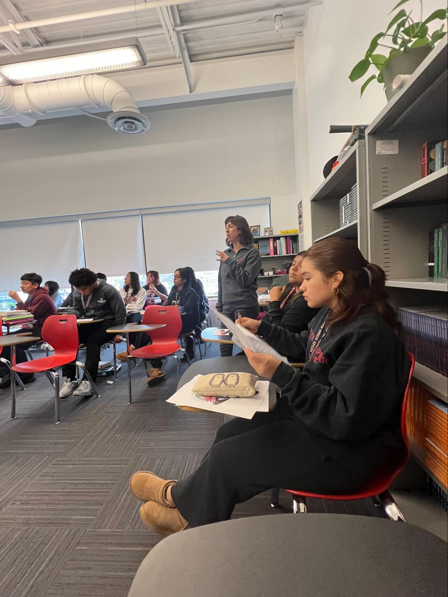 This is Ms. Swain's AP English class she is standing at the side of the room talking to students while they are reading along with a worksheet. The students are sitting at their desk alined along the classroom.