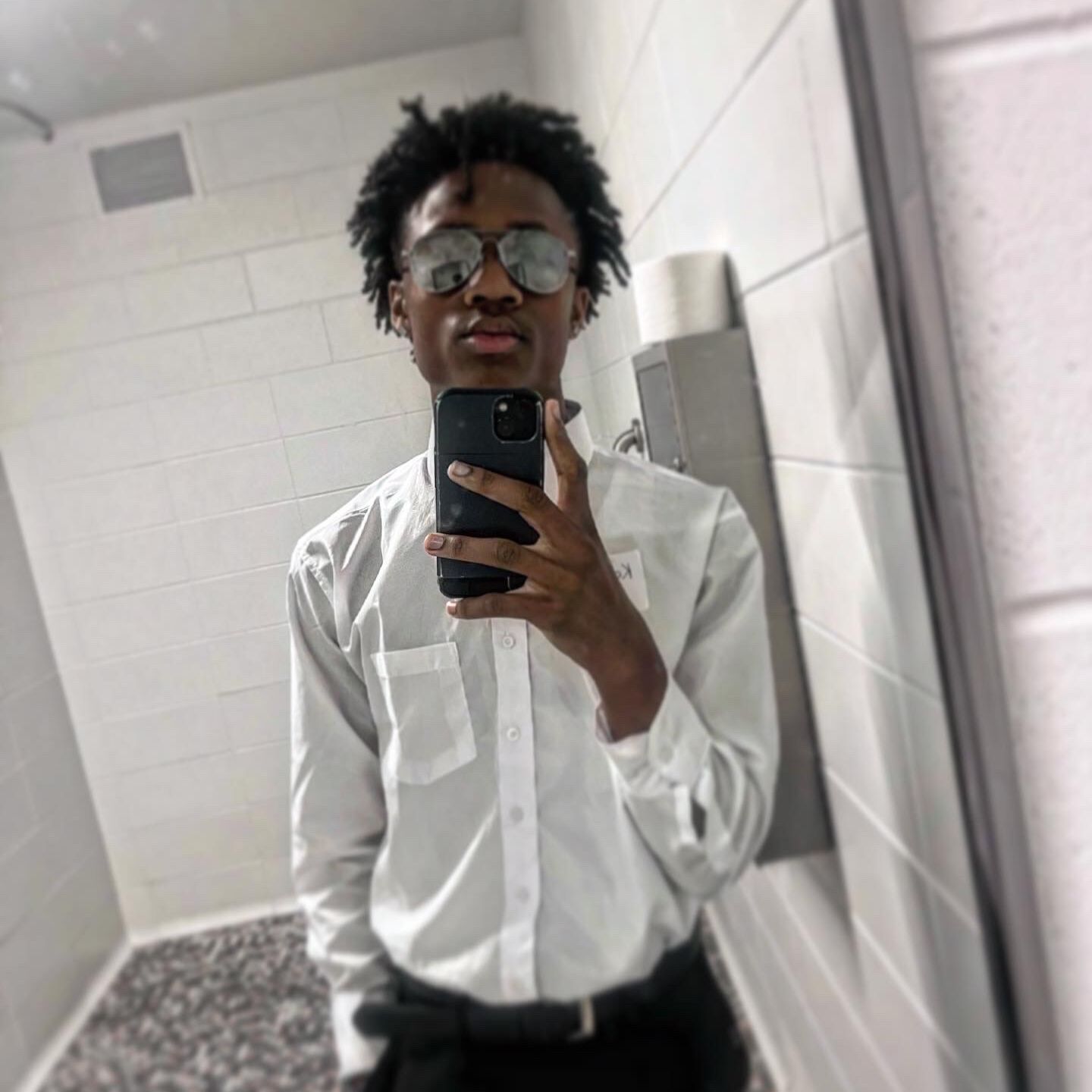 In this photo, Kamauri I, a senior at Chicago Bulls College Prep, is taking a selfie in a mirror. He is a young Black man with short black hair, wearing shades, a white button-up shirt and black dress pants.