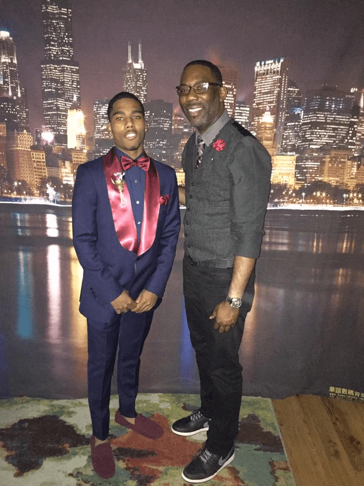 In this photo, Carlos Duncan stands with one of his students who is dressed up in a gorgeous navy suit with red lapels and a red bowtie. They are standing in front of Lake Michigan with the Chicago downtown skyline behind them.