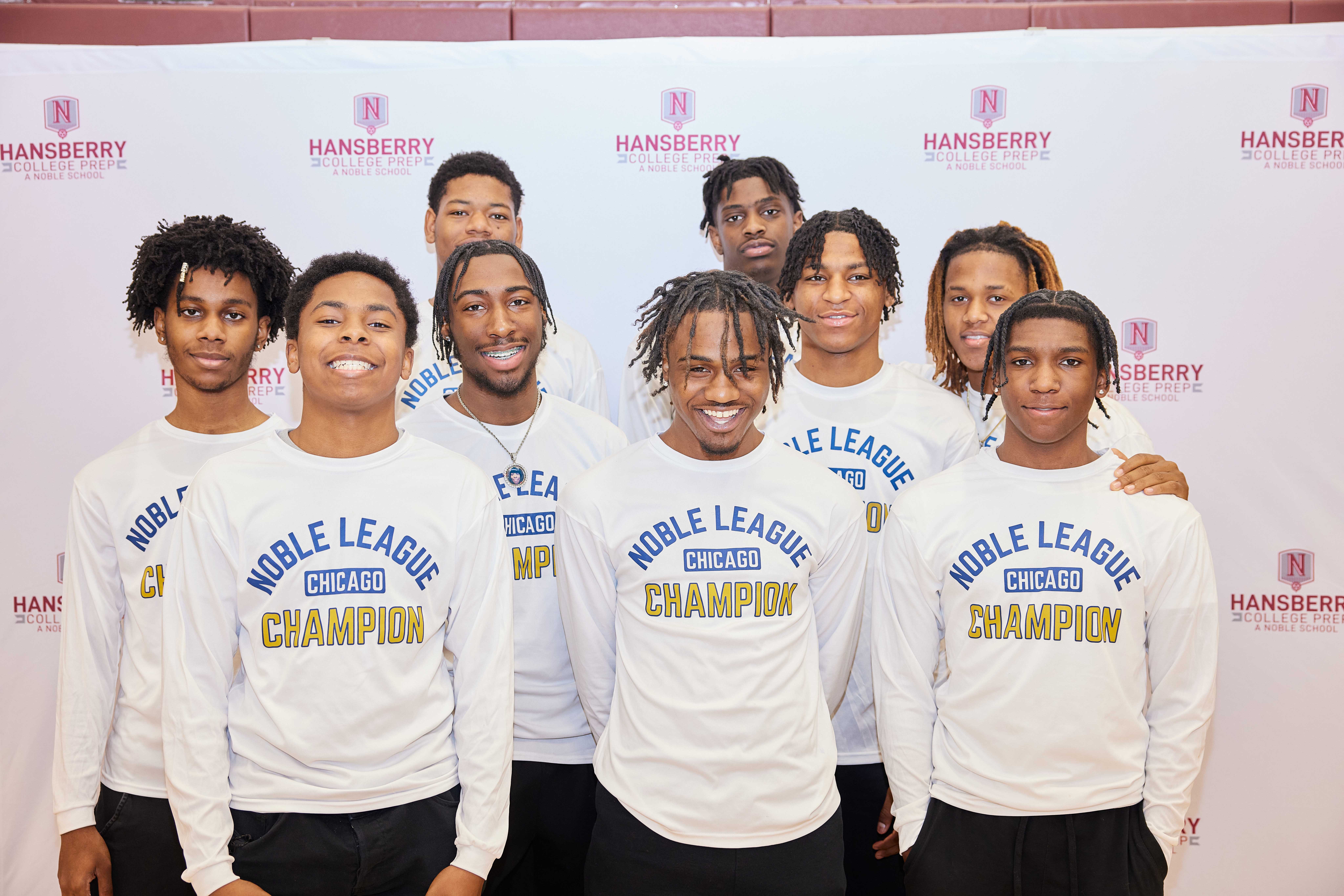 This photo shows the Hansberry College Prep boys' varsity basketball team all posing and smiling in front of a white backdrop with the HCP logo repeated on it.