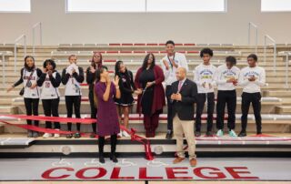 This photo shows a group of Hansberry student-athletes with Noble Schools CEO Constance Jones, Principal Kashawndra Wilson, and Illinois State Representative Justin Slaughter standing on the bleachers of the new Hansberry gym renovation, smiling and clapping after cutting the ribbon at the ribbon-cutting ceremony. Principal Wilson holds the large pair of shiny gold scissors that were used to cut the ribbon, which is now lying on the floor.