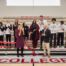 This photo shows a group of Hansberry student-athletes with Noble Schools CEO Constance Jones, Principal Kashawndra Wilson, and Illinois State Representative Justin Slaughter standing on the bleachers of the new Hansberry gym renovation, smiling and clapping after cutting the ribbon at the ribbon-cutting ceremony. Principal Wilson holds the large pair of shiny gold scissors that were used to cut the ribbon, which is now lying on the floor.