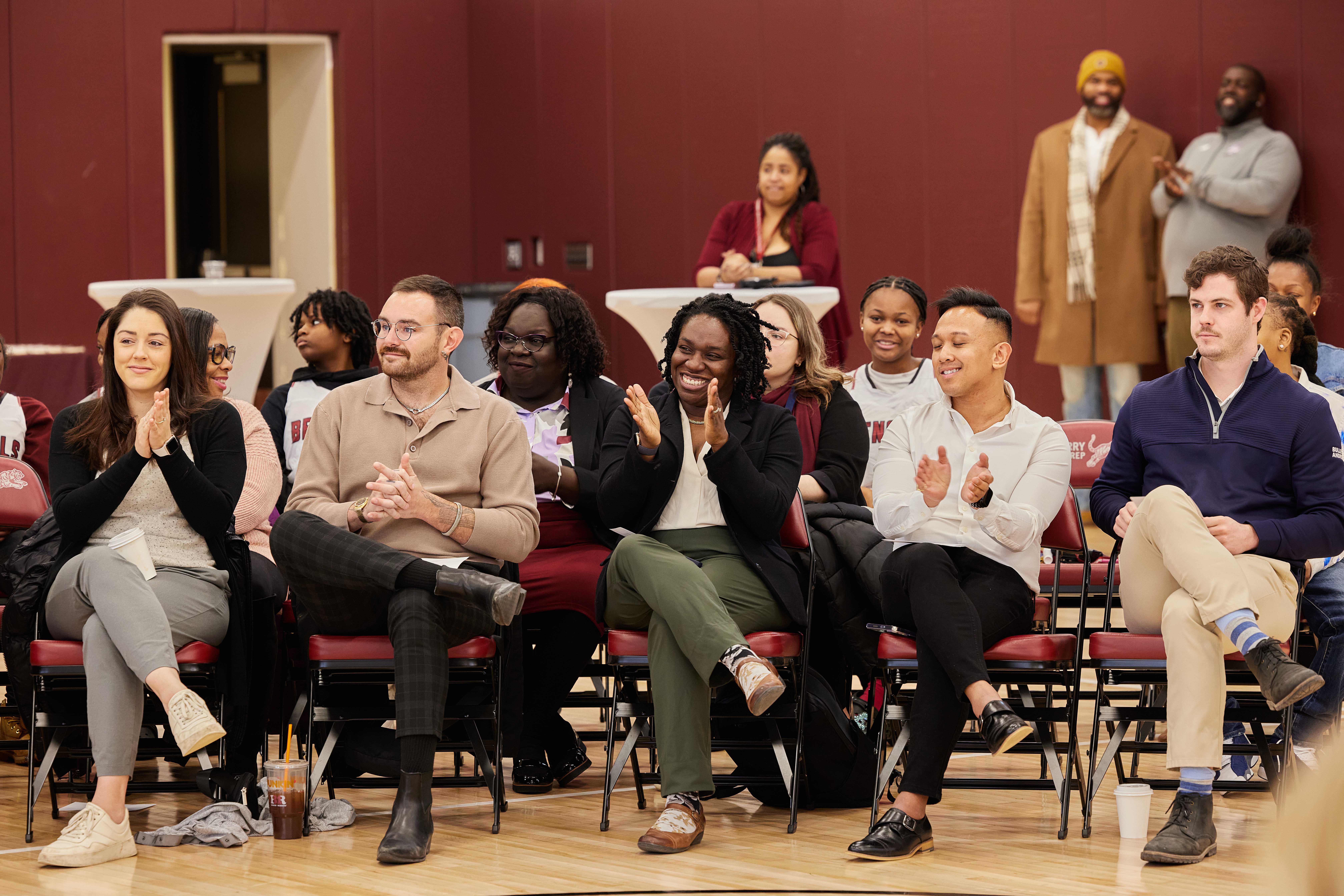 This photo shows a crowd of people sitting in chairs on a gym floor clapping and looking in the same direction at the Hansberry College Prep ribbon-cutting ceremony for their new gym renovation.